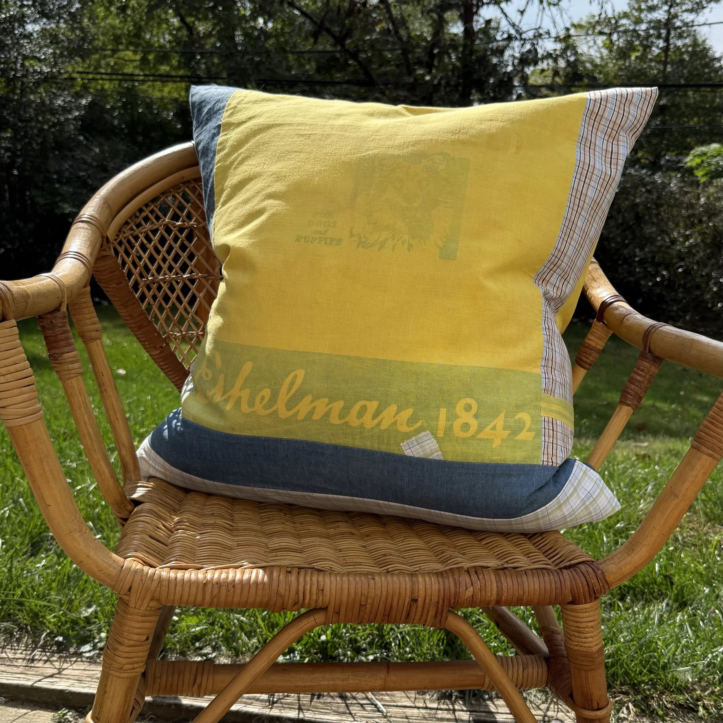 A wicker chair with a yellow cushion that has a design and the words 'Shelman 1842' on it, placed outdoors on grass with trees in the background.