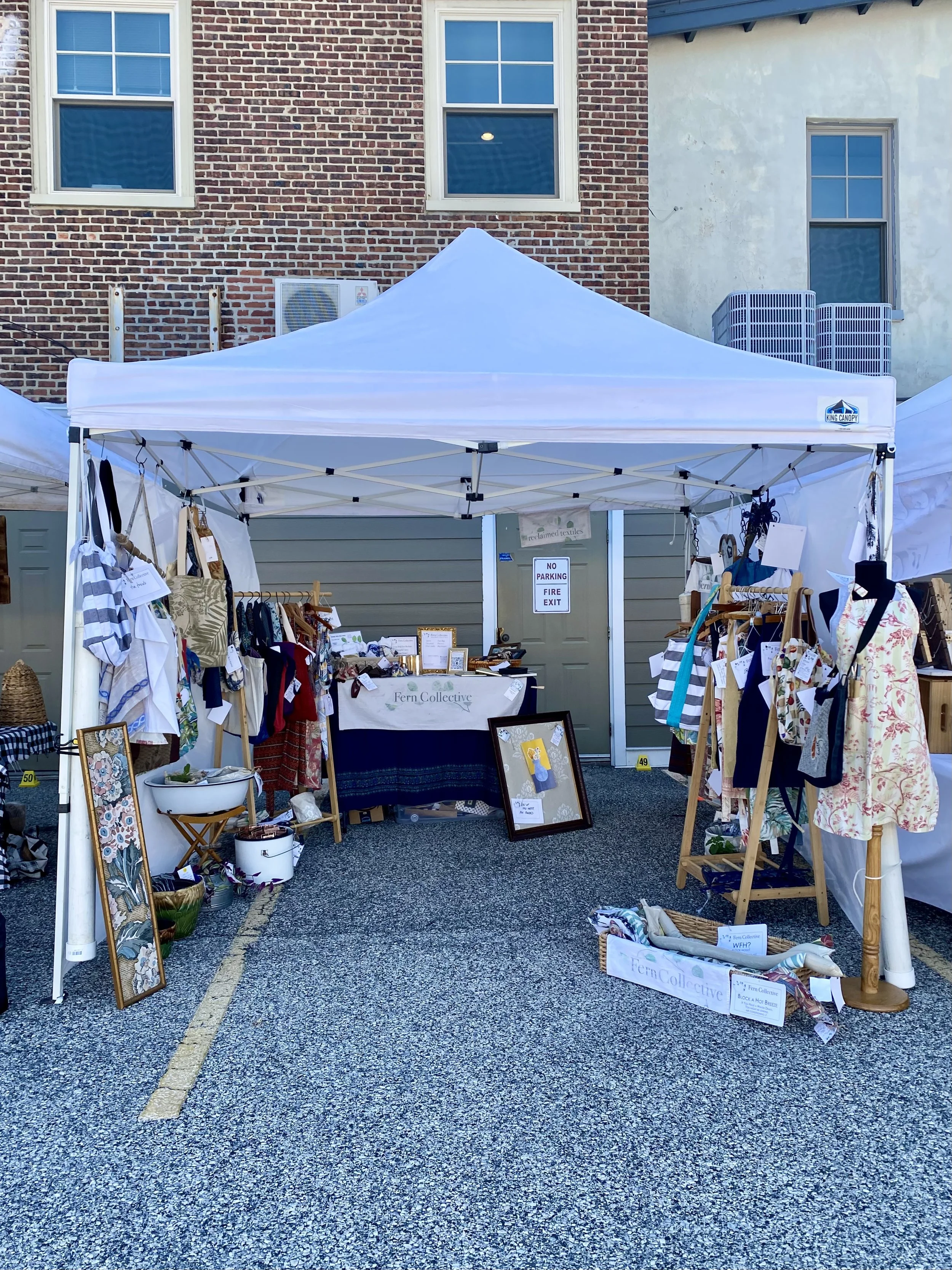 Outdoor craft fair booth with handmade textiles, bags, and art displayed under a white canopy tent outside against brick and stucco walls.