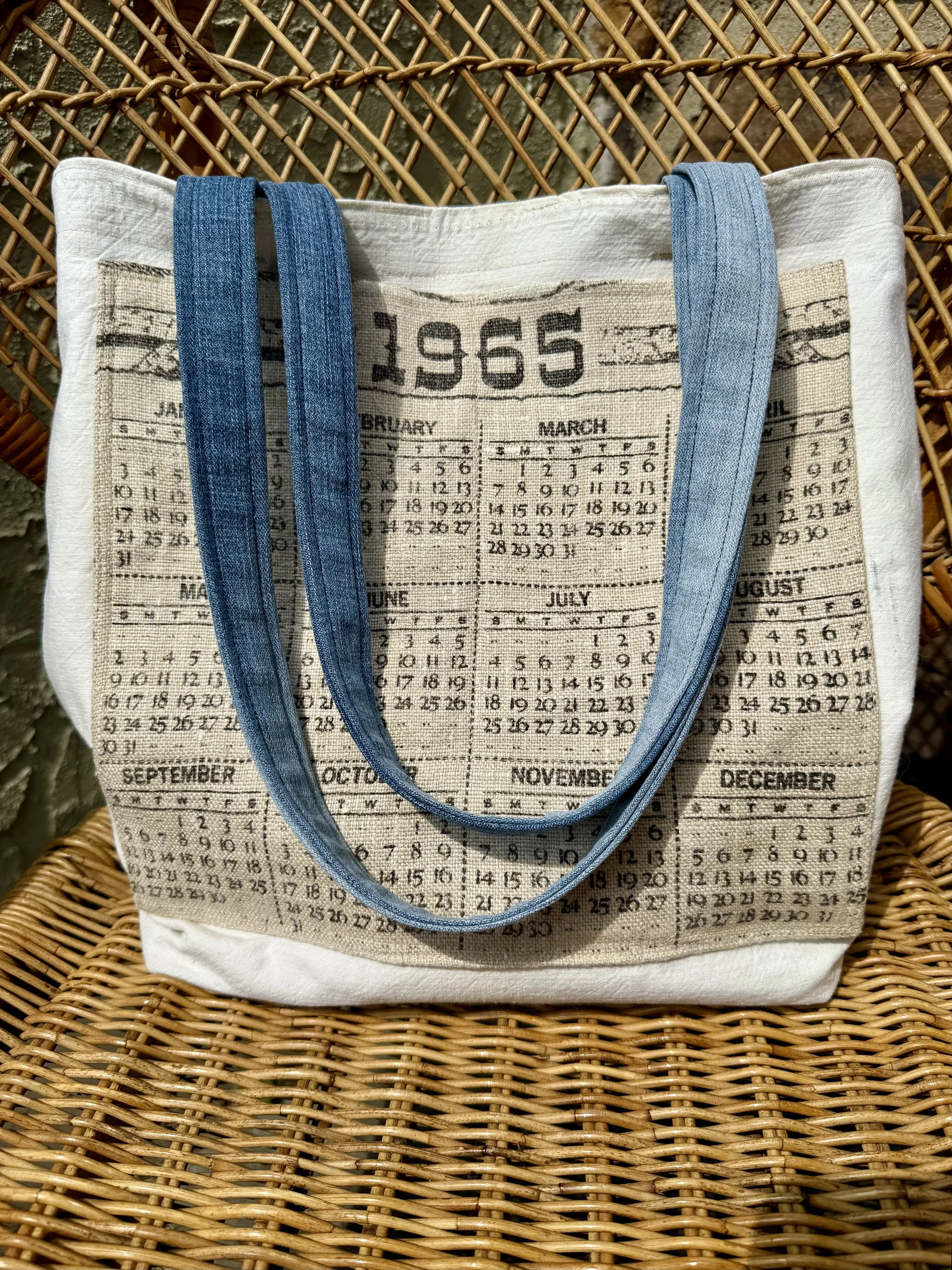 Linen tote bag with a vintage calendar print and blue denim-style straps, placed on a wicker chair.