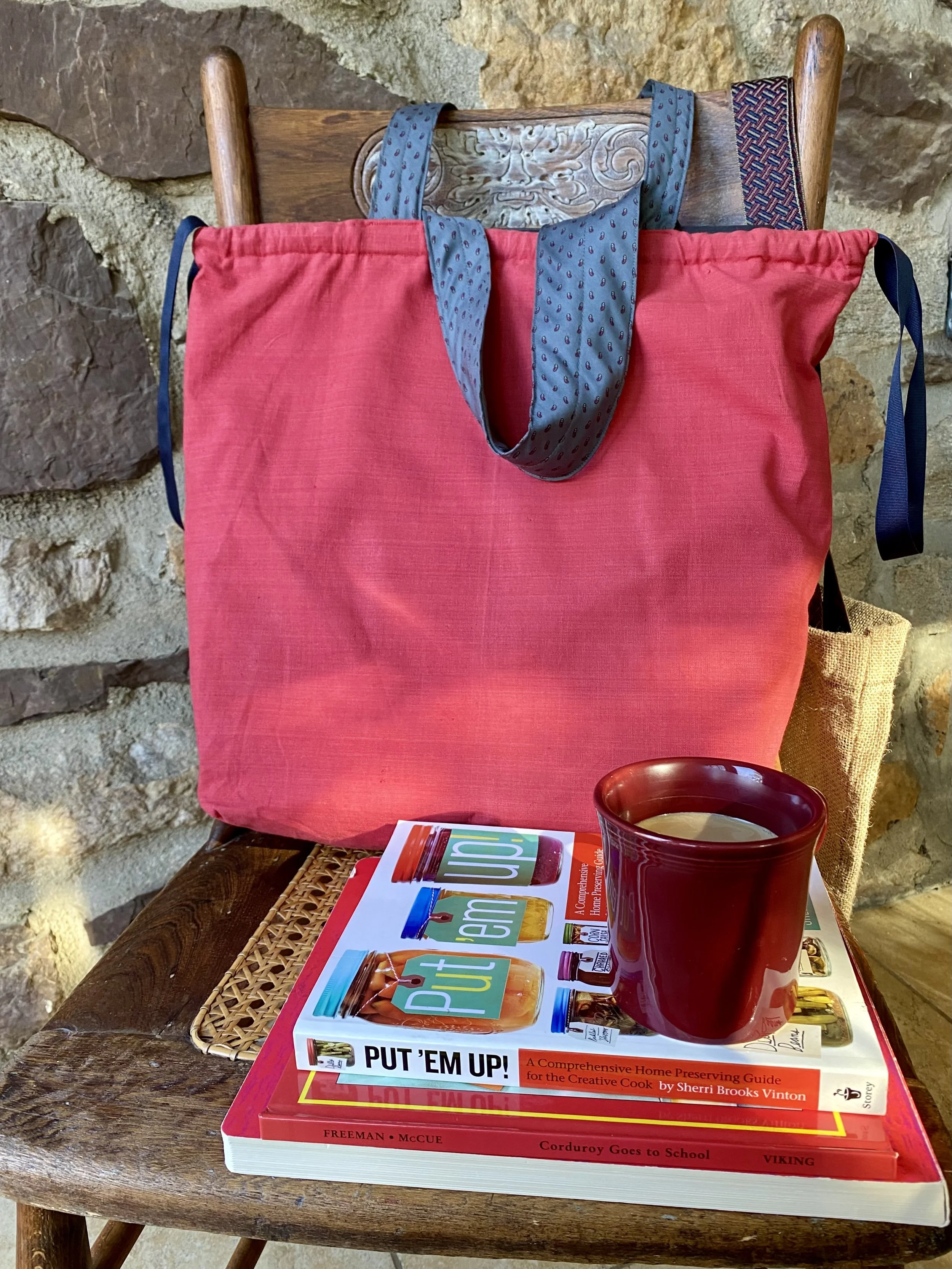 A wooden chair with a stone wall background. On the chair, a large pink tote bag with a blue patterned scarf draped over the top. On the table in front of the chair, there are two colorful cookbooks, a red mug filled with a hot beverage, and a small 