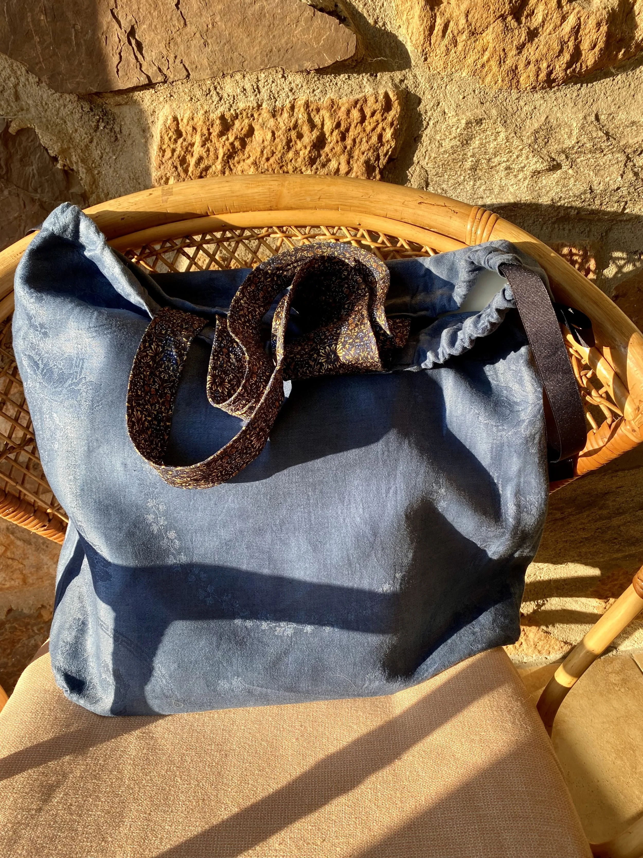 Blue tote bag with a patterned fabric handle on a rattan chair with sunlight and shadows, against a stone wall background.