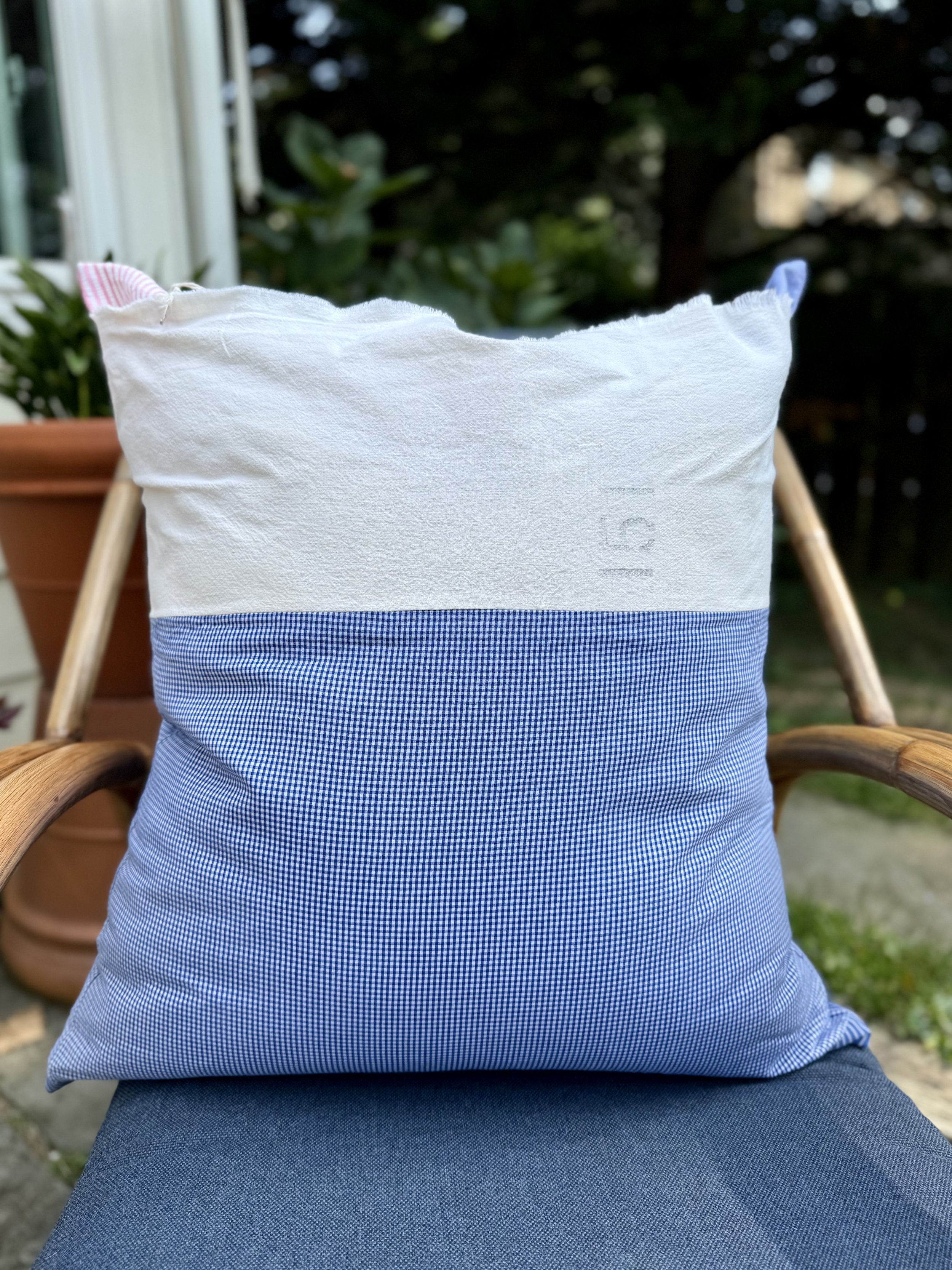 A pillow with a blue and white gingham pattern on the bottom half and plain white on the top half, placed on a chair outdoors during daytime.