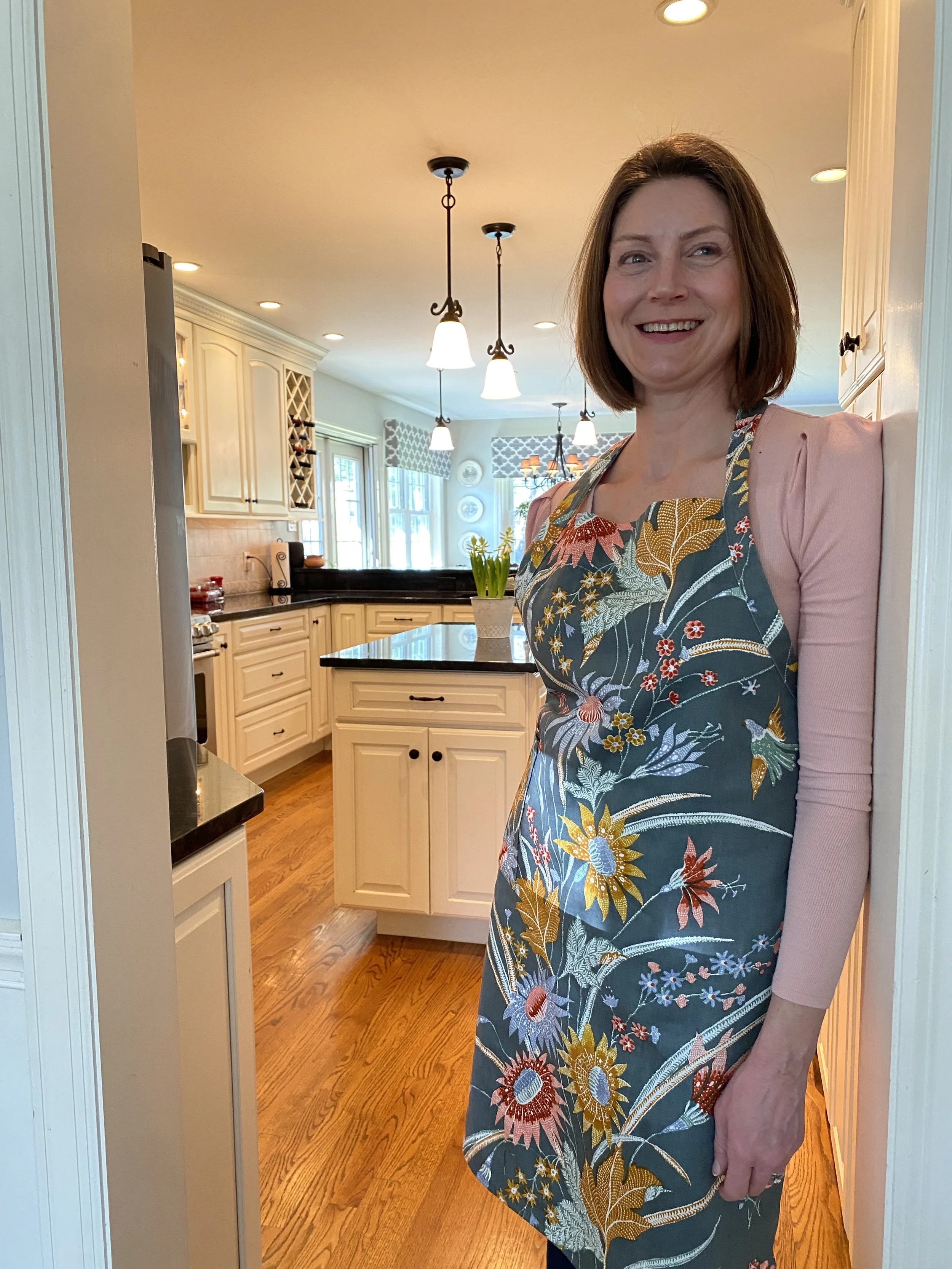 A woman smiles in a kitchen, standing near a doorway, wearing a floral apron over a pink long-sleeve shirt.