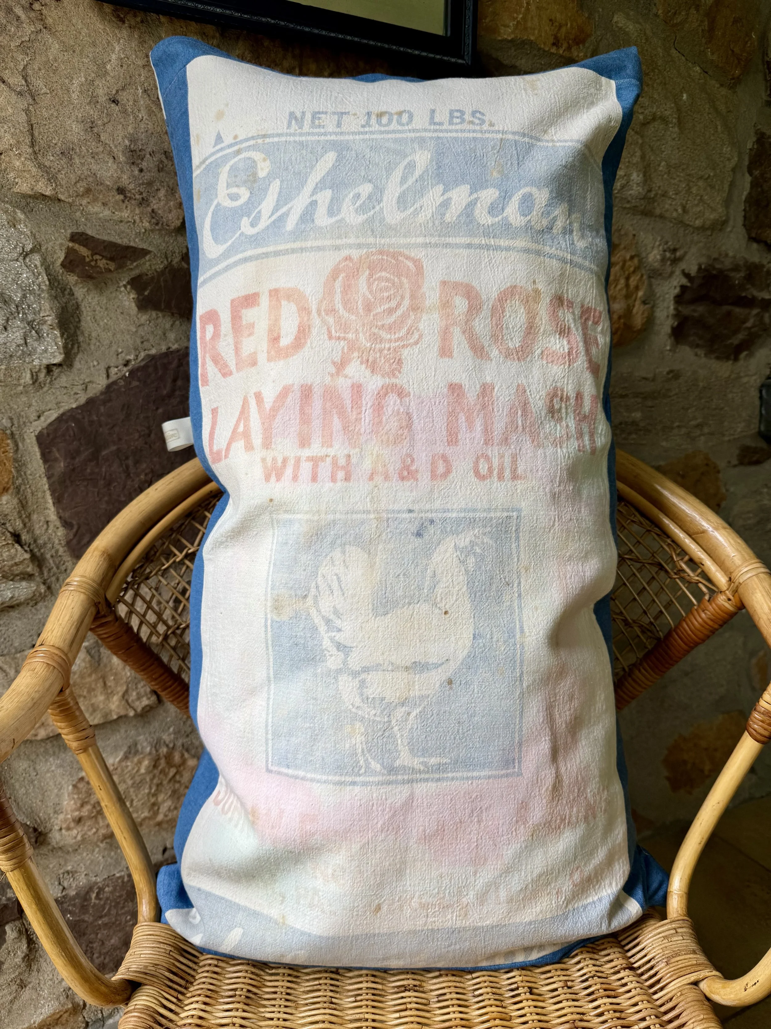 Decorative pillow shaped like a bag with vintage-style advertising for Red Rose and chocolate, placed on a wicker chair against a stone wall.