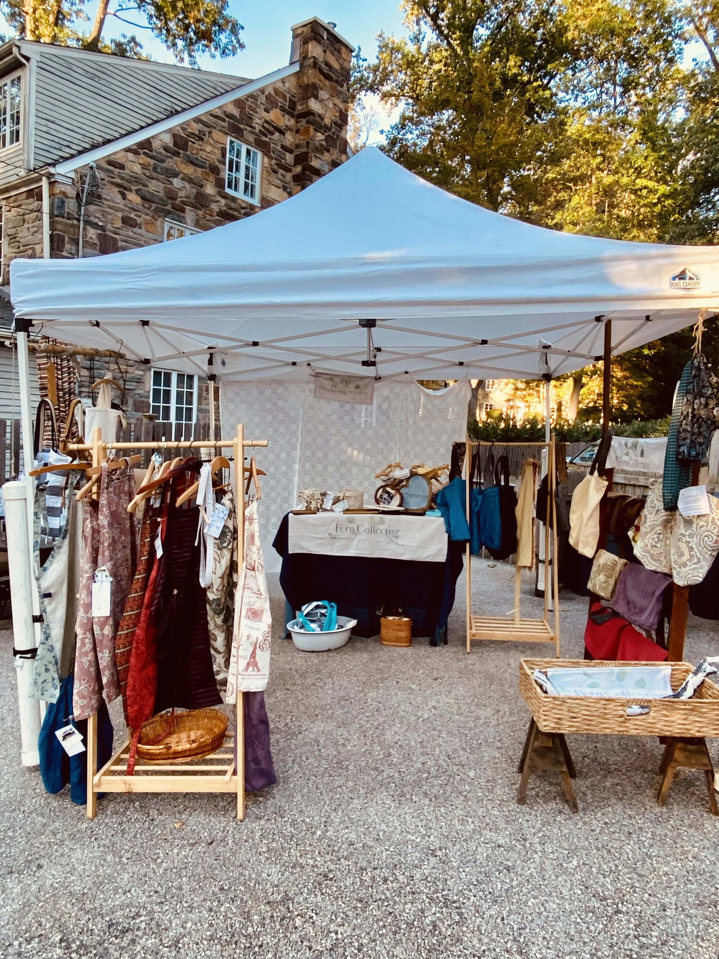 Outdoor craft fair booth with clothing, bags, and accessories under a white canopy on a sunny day.