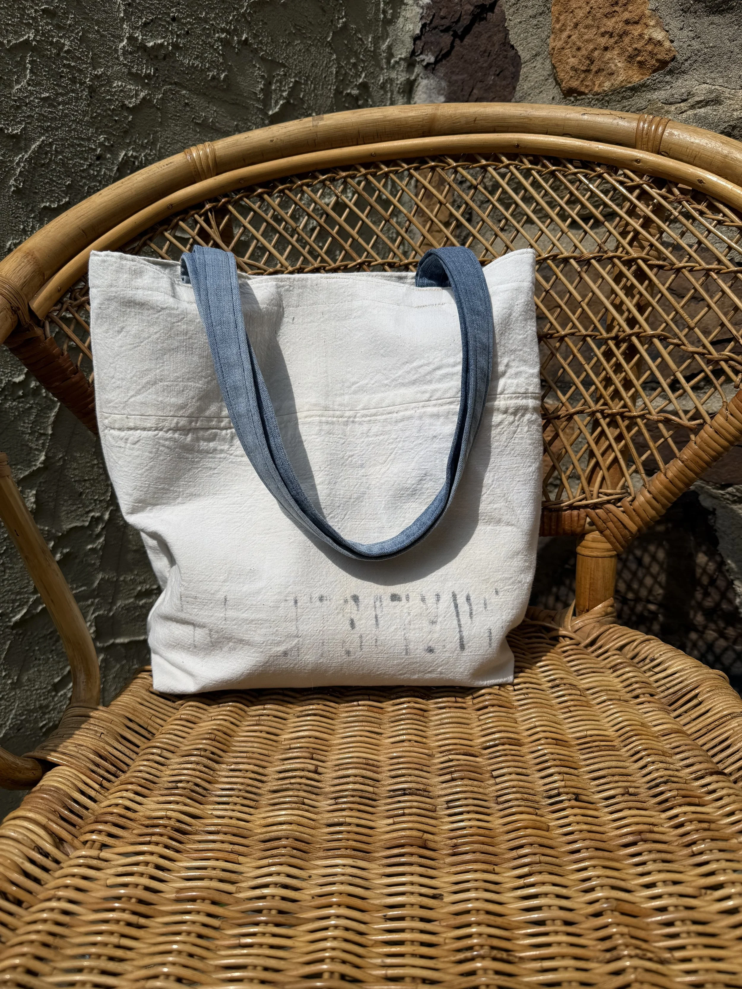 White tote bag with denim straps resting on a wicker chair, with a textured stone wall in the background.