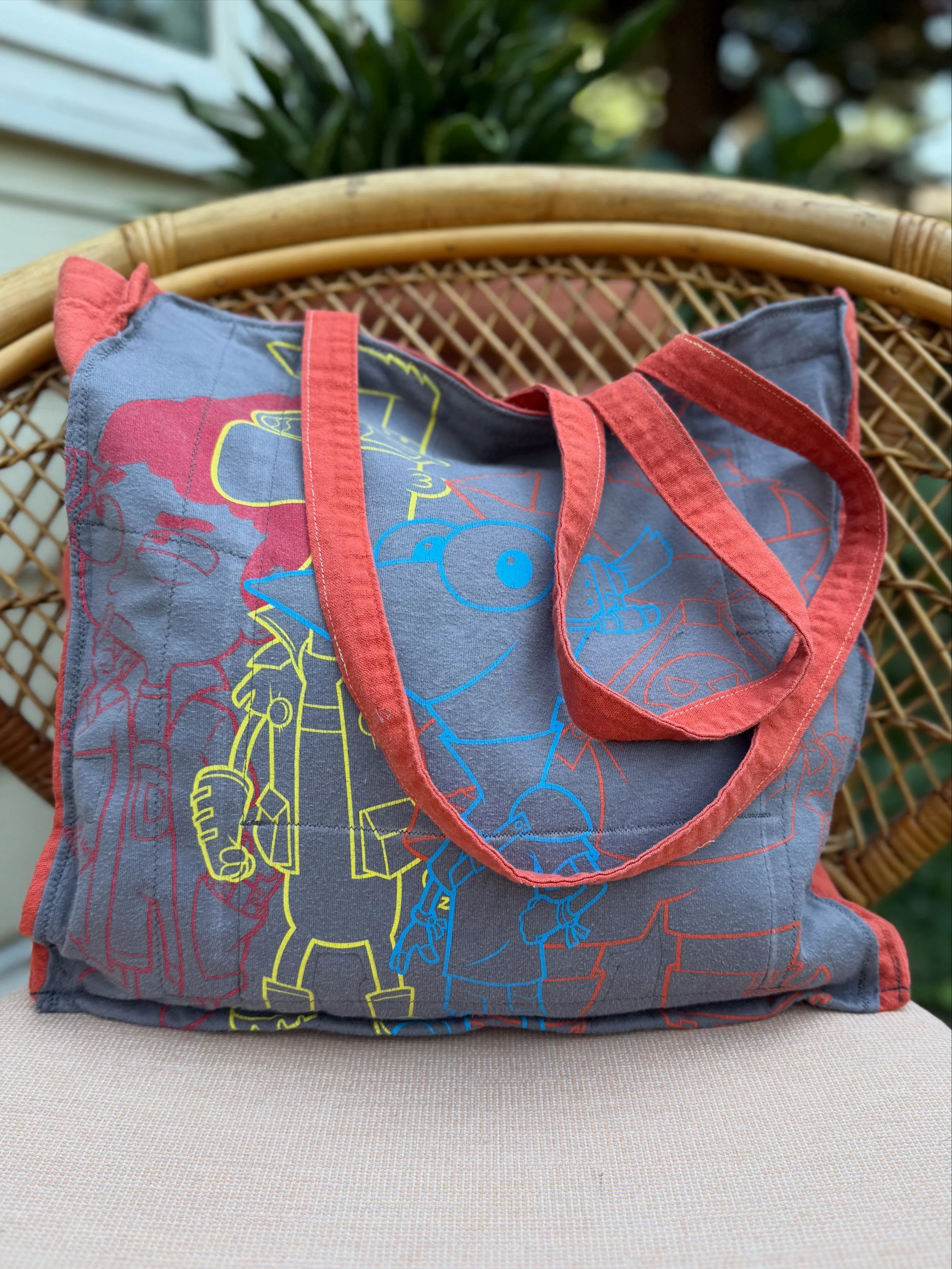 Gray tote bag with colorful Cartoon Network characters and red handles on a chair outside.