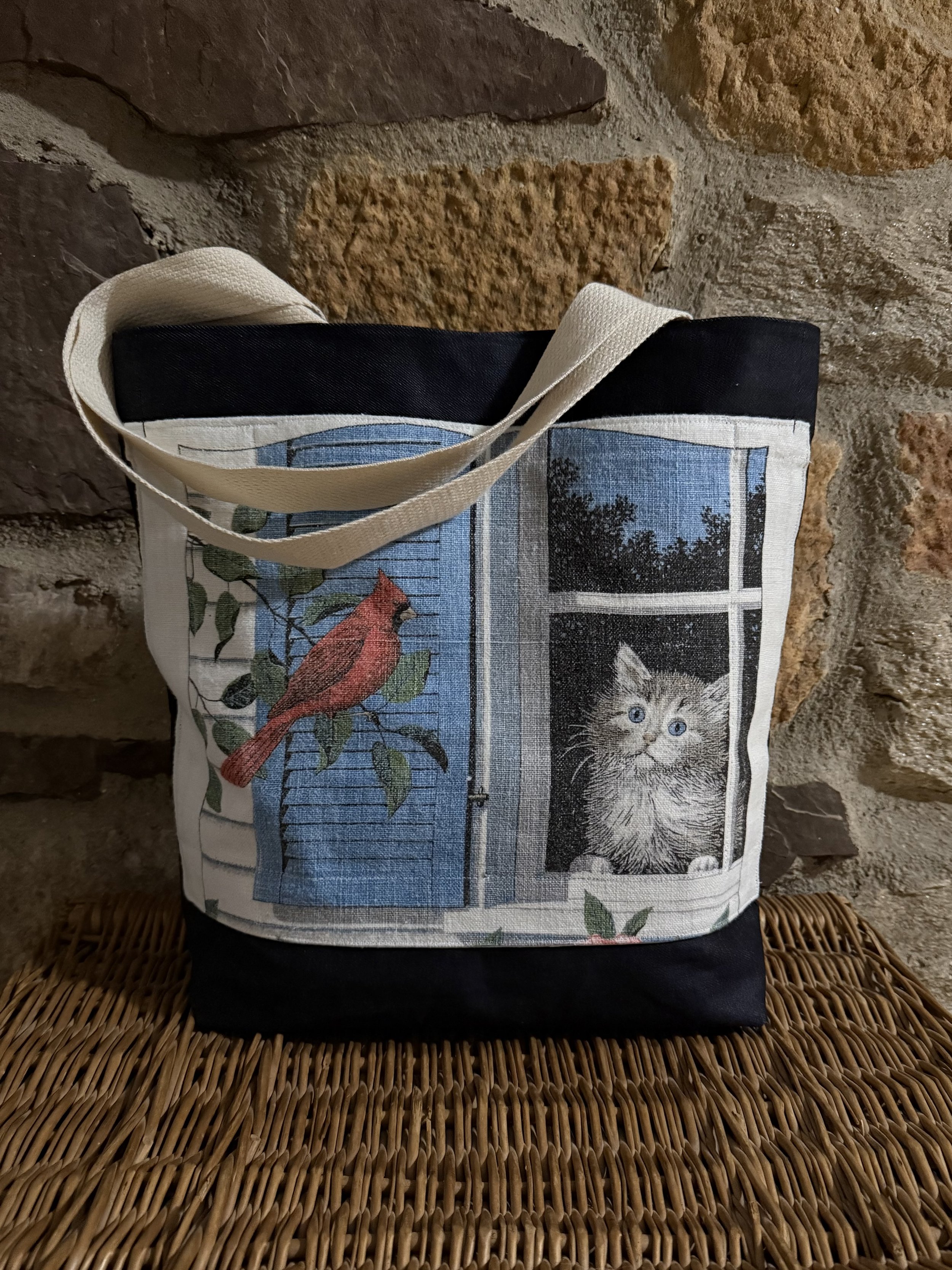 A canvas tote bag with a print of a cat looking out a window and a cardinal bird on a branch, placed on a wicker surface against a brick wall background.