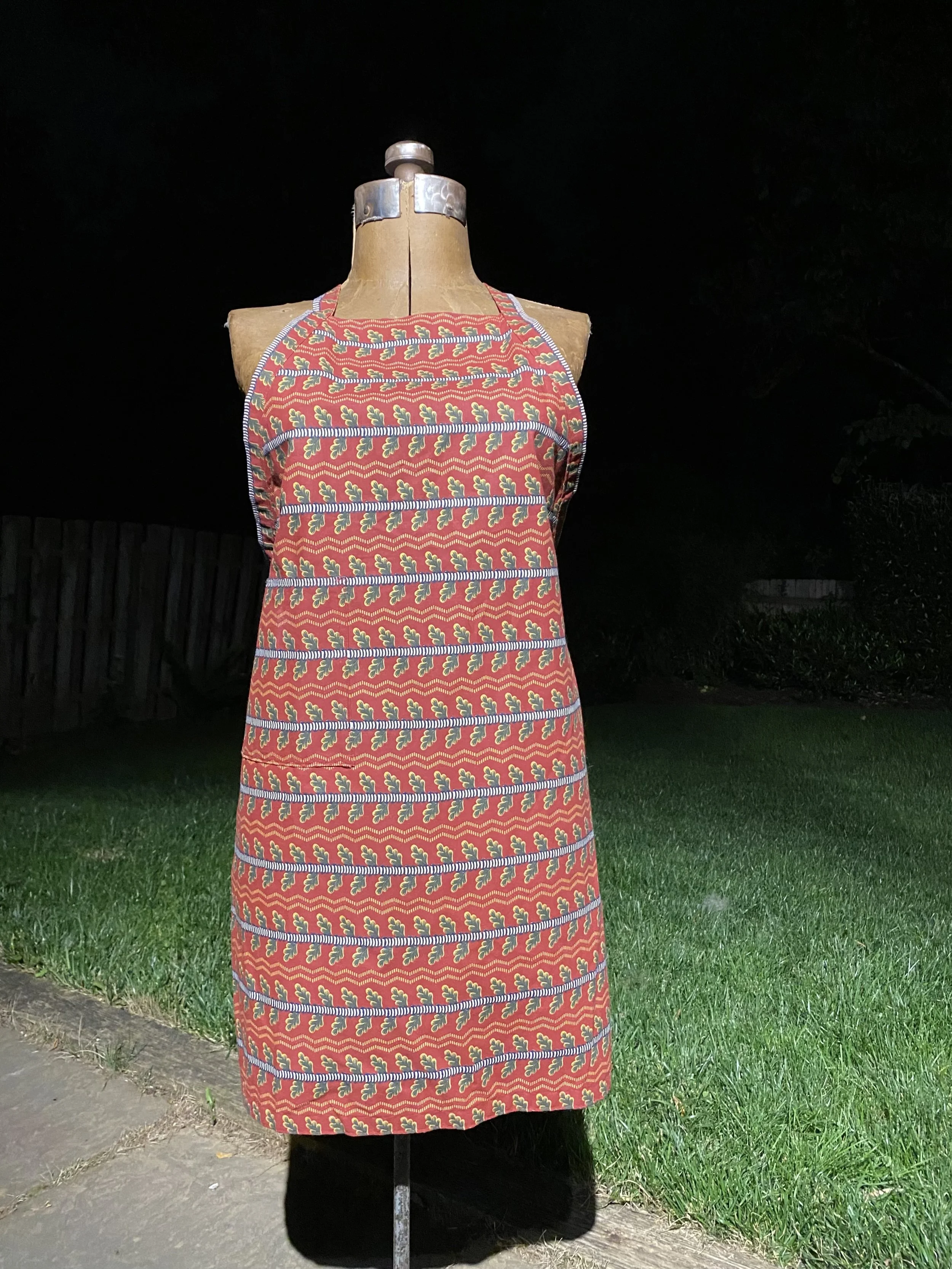 A mannequin wearing a sleeveless, knee-length garment with a red background and a repeating pattern of green and yellow leaves, displayed outdoors at night.