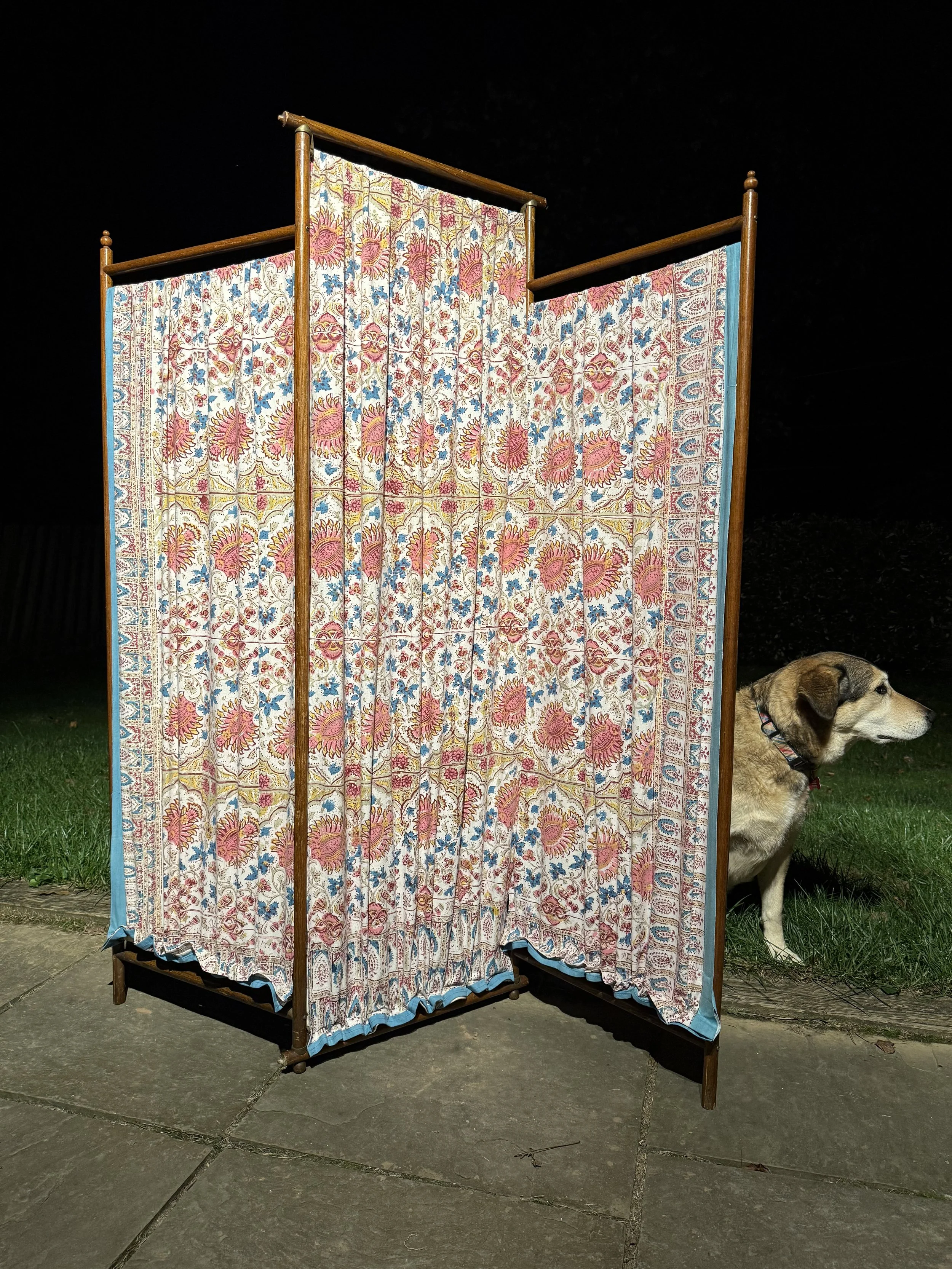 A wooden room divider with floral-patterned fabric panels standing outdoors at night, next to a dog sitting on grass.
