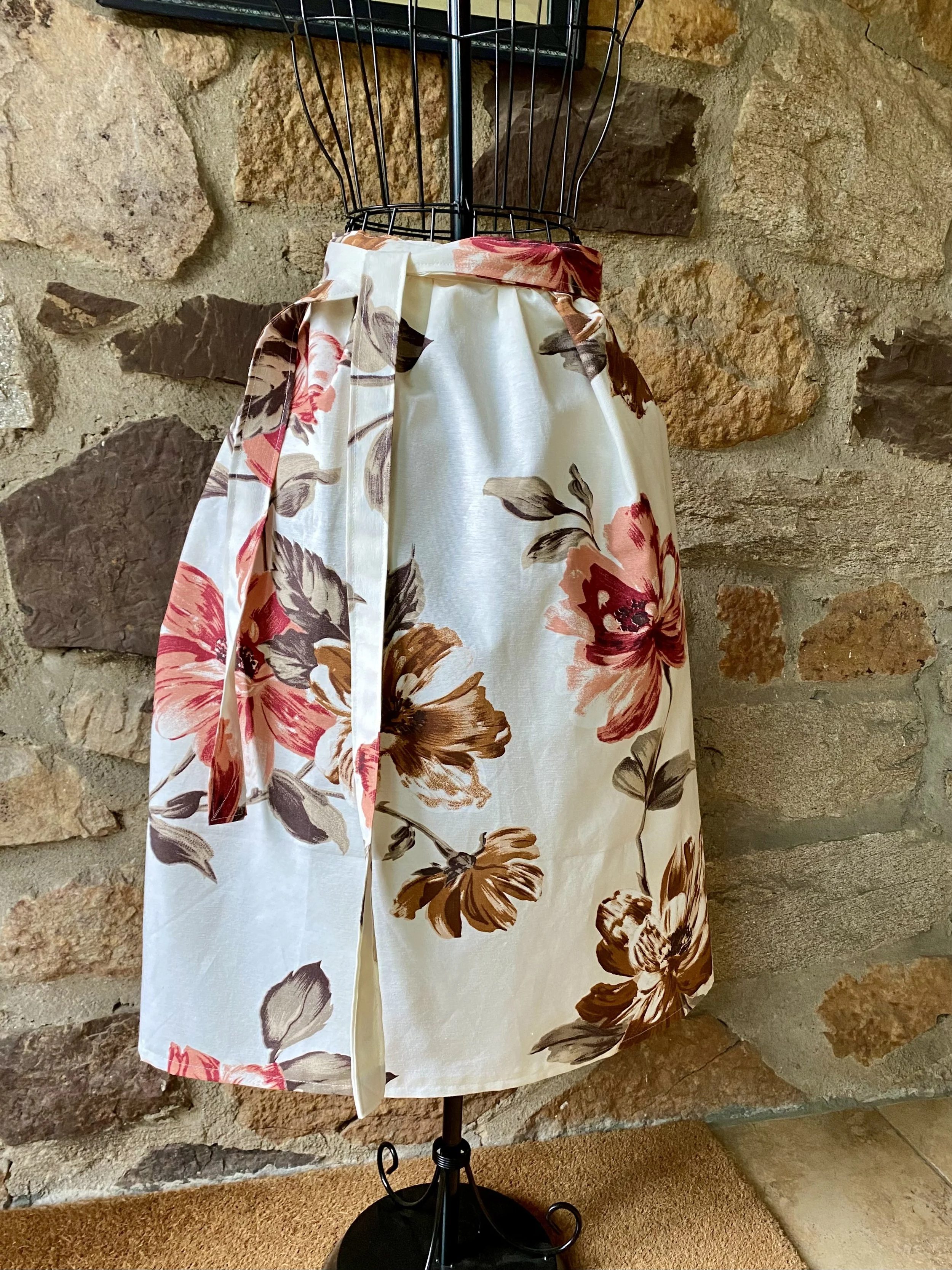 A floral skirt with pink, brown, and gray flowers on a cream background, displayed on a black dress form against a stone wall.