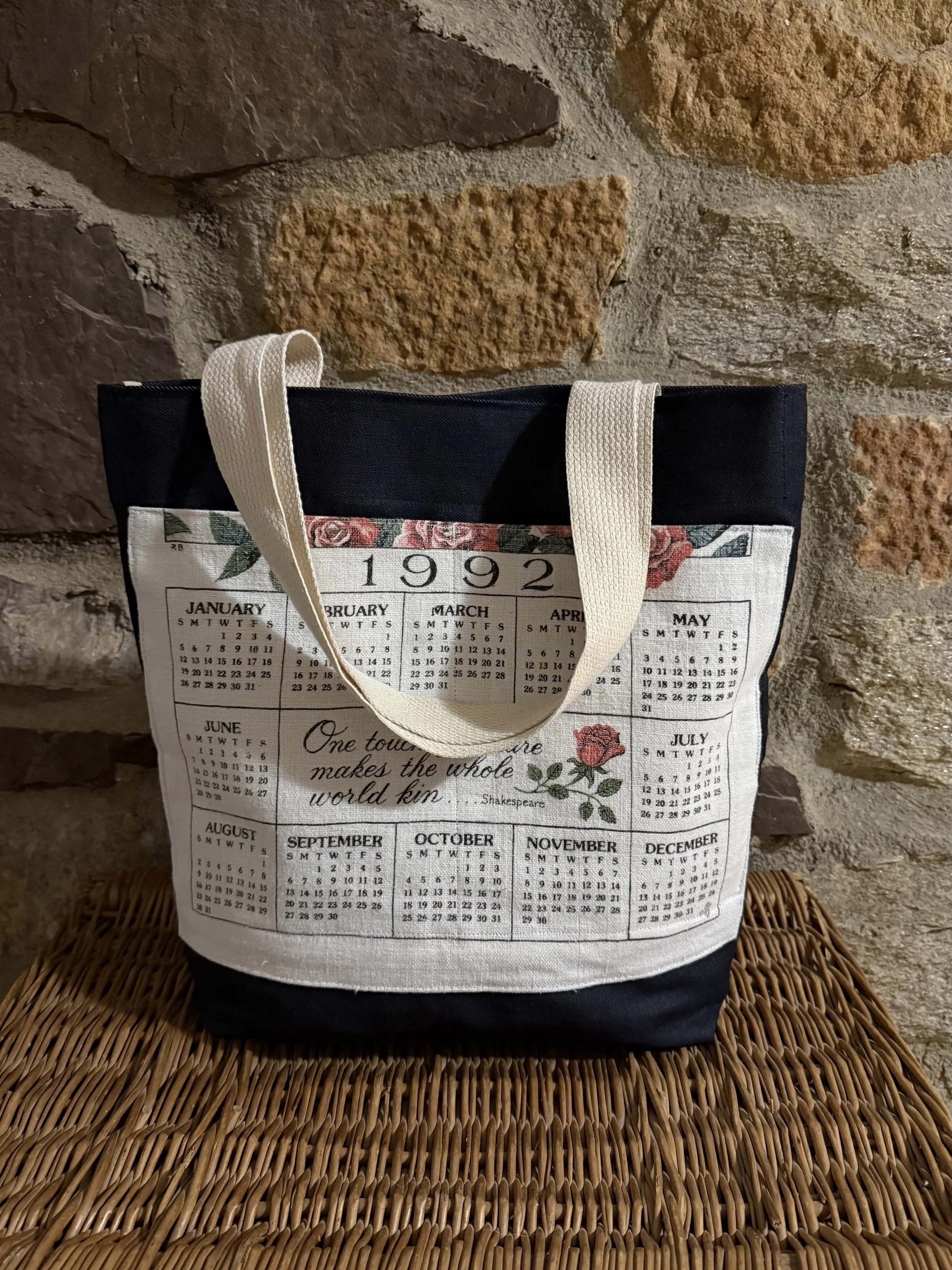A tote bag with a vintage-style 1992 calendar design, featuring months, days, a quote by Shakespeare, and a rose illustration, placed on a wicker surface against a stone brick wall.