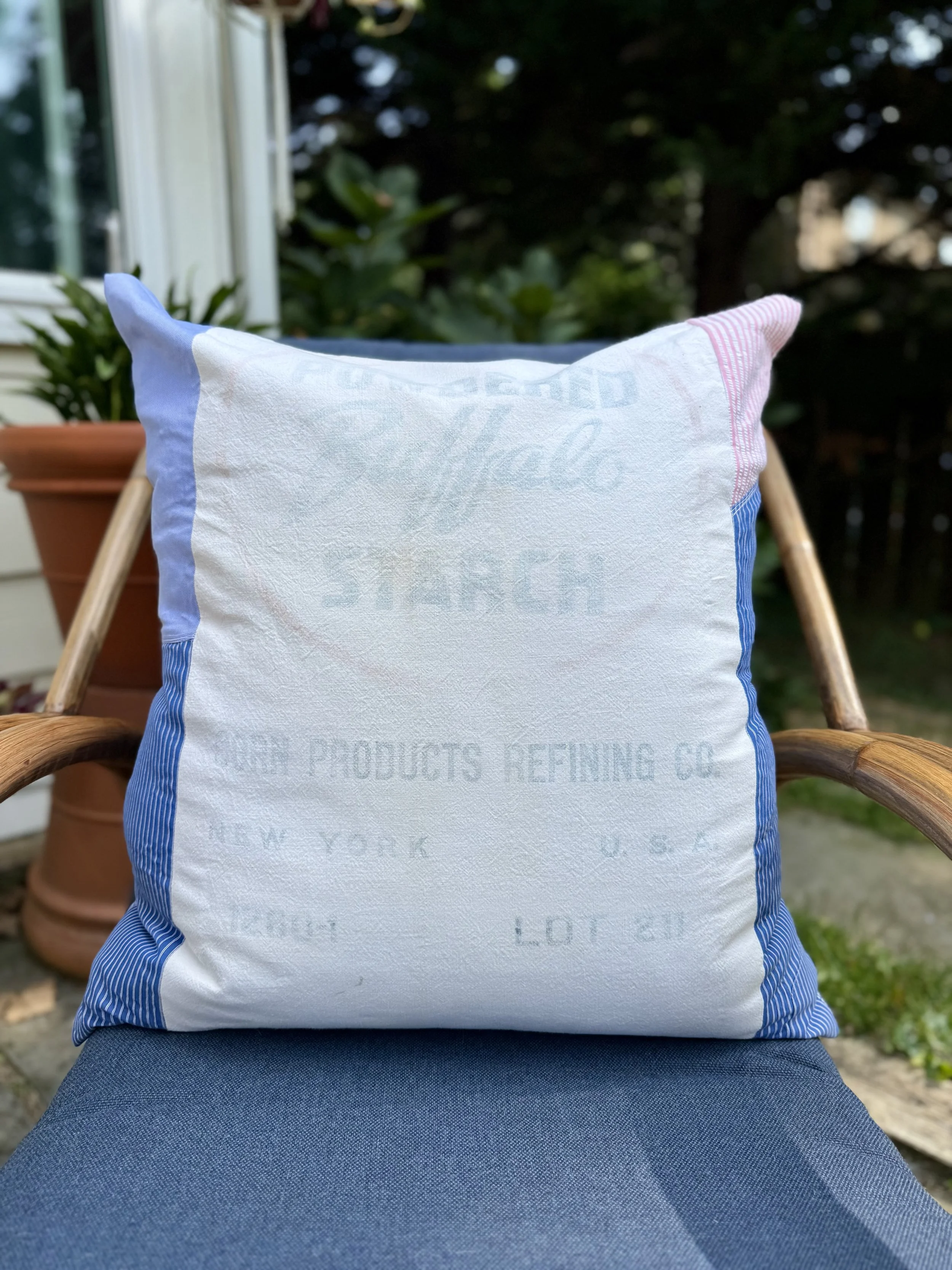 A decorative pillow with vintage-style printing, resting on a dark blue outdoor chair, with potted plants and greenery in the background.