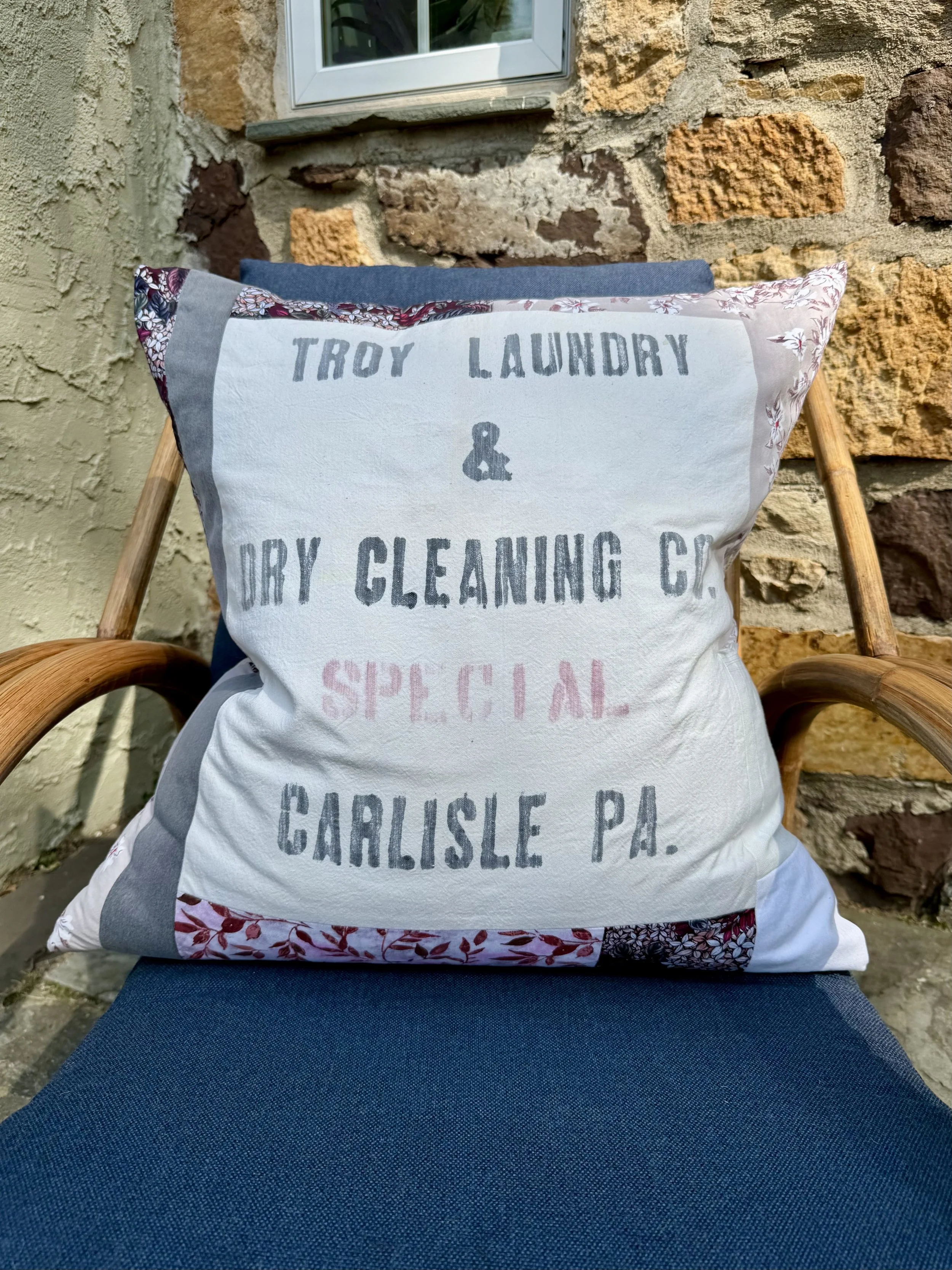 A decorative pillow with text that reads 'Troy Laundry & Dry Cleaning Co. Special Carlisle PA,' resting on a blue outdoor chair with wooden armrests, against a brick and stucco wall.