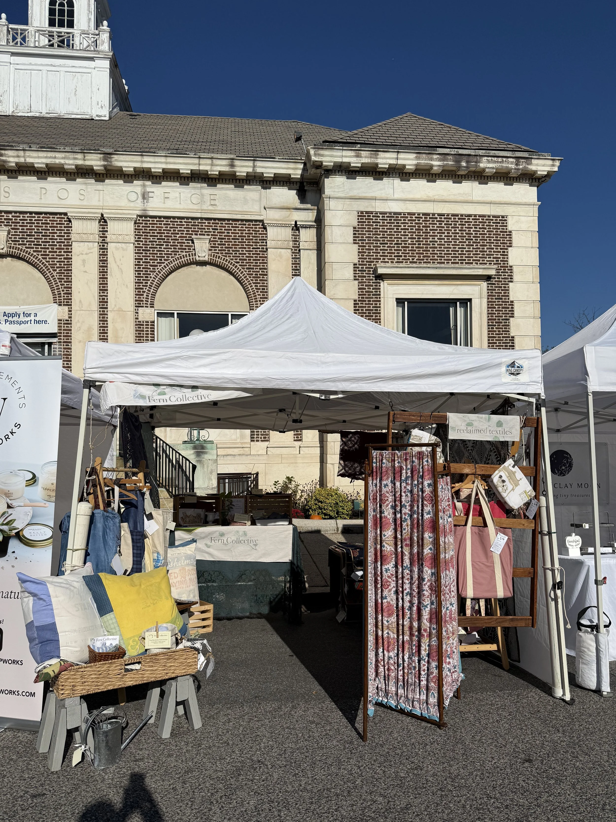 Outdoor market stall with textiles, pillows, and accessories set up in front of a historic brick and stone building, under white canopy tents on a sunny day.