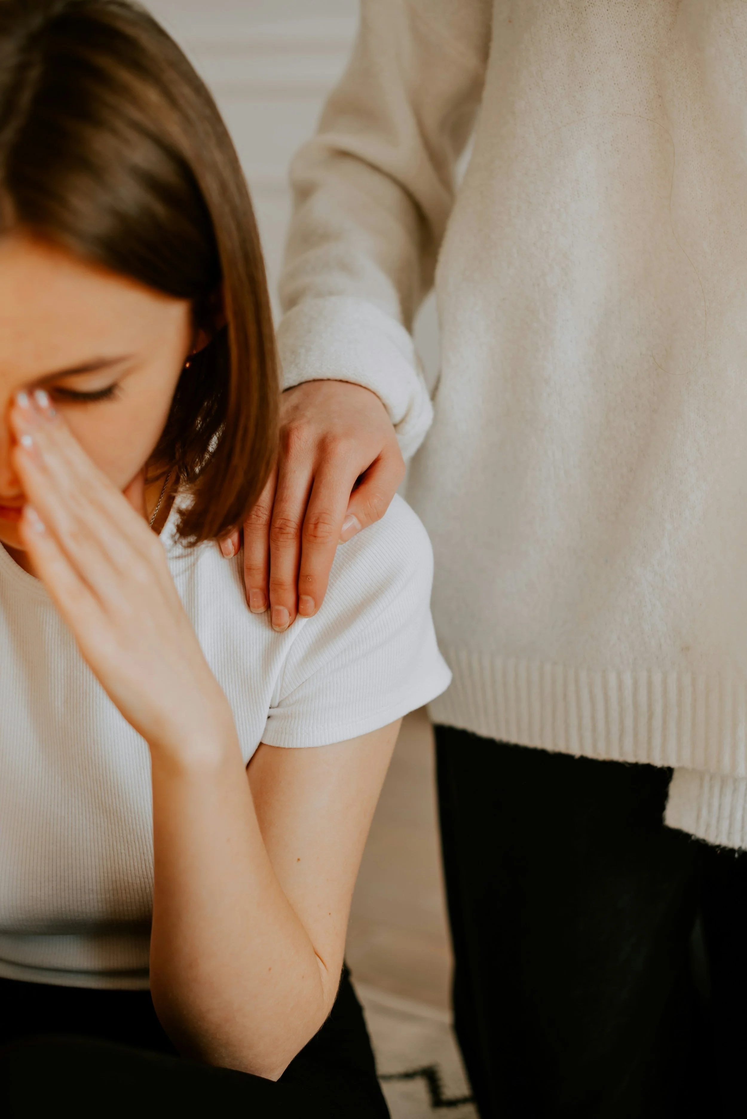 A person with brown hair appears distressed, holding her forehead with her left hand. Another person, standing beside her, places a comforting hand on her shoulder.