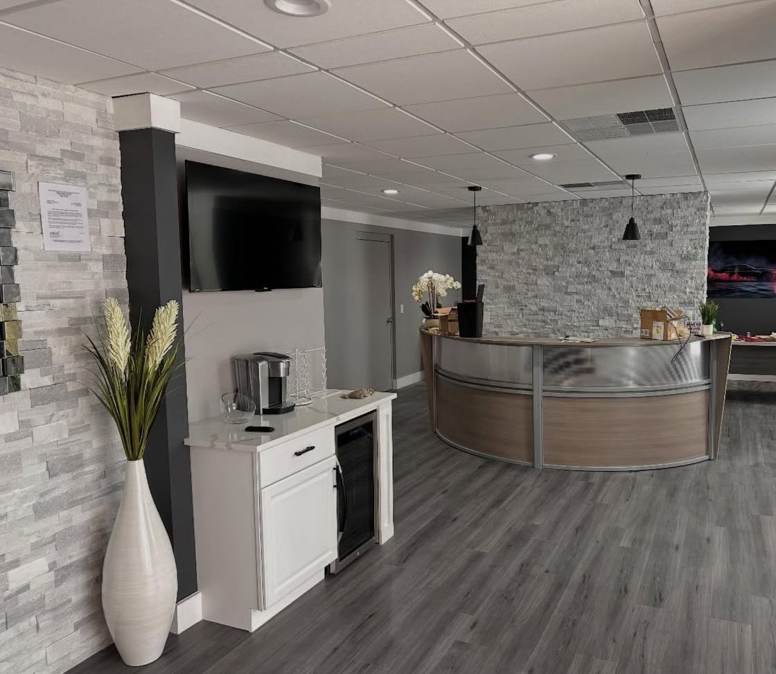 Modern lobby area with a curved reception desk, wall-mounted TV, potted plants, a small white cabinet with a coffee maker, and gray wood-like flooring.