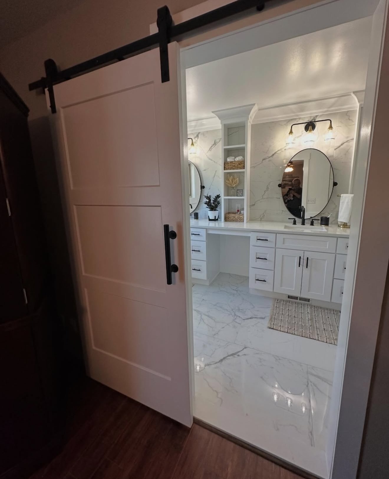 View of a modern bathroom with white cabinetry, marble countertops, and round mirrors, seen through a sliding barn door.