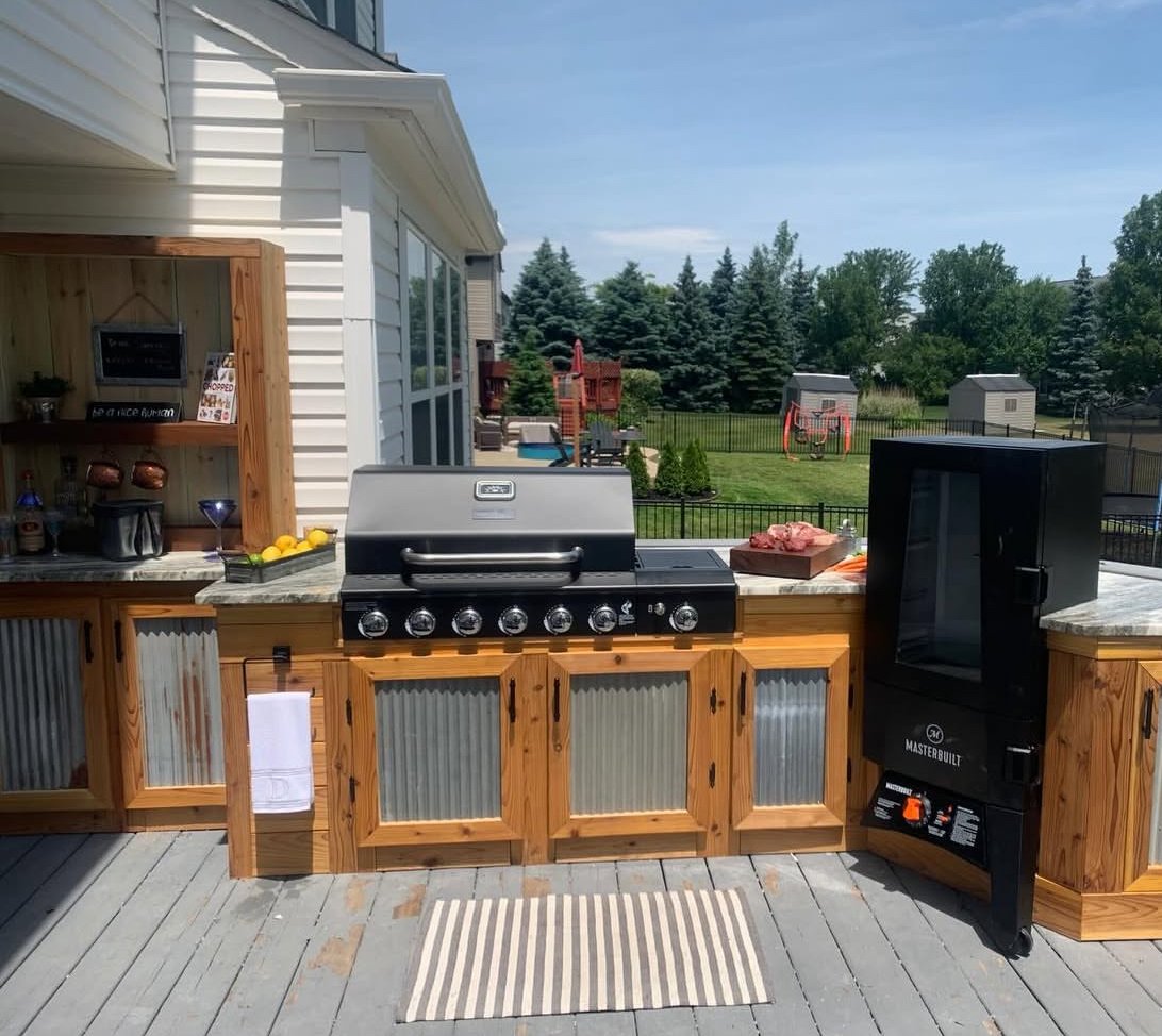 Outdoor kitchen with a grill, black smoker, and wooden cabinets on a deck, overlooking a backyard with a playset, trees, and a fence.