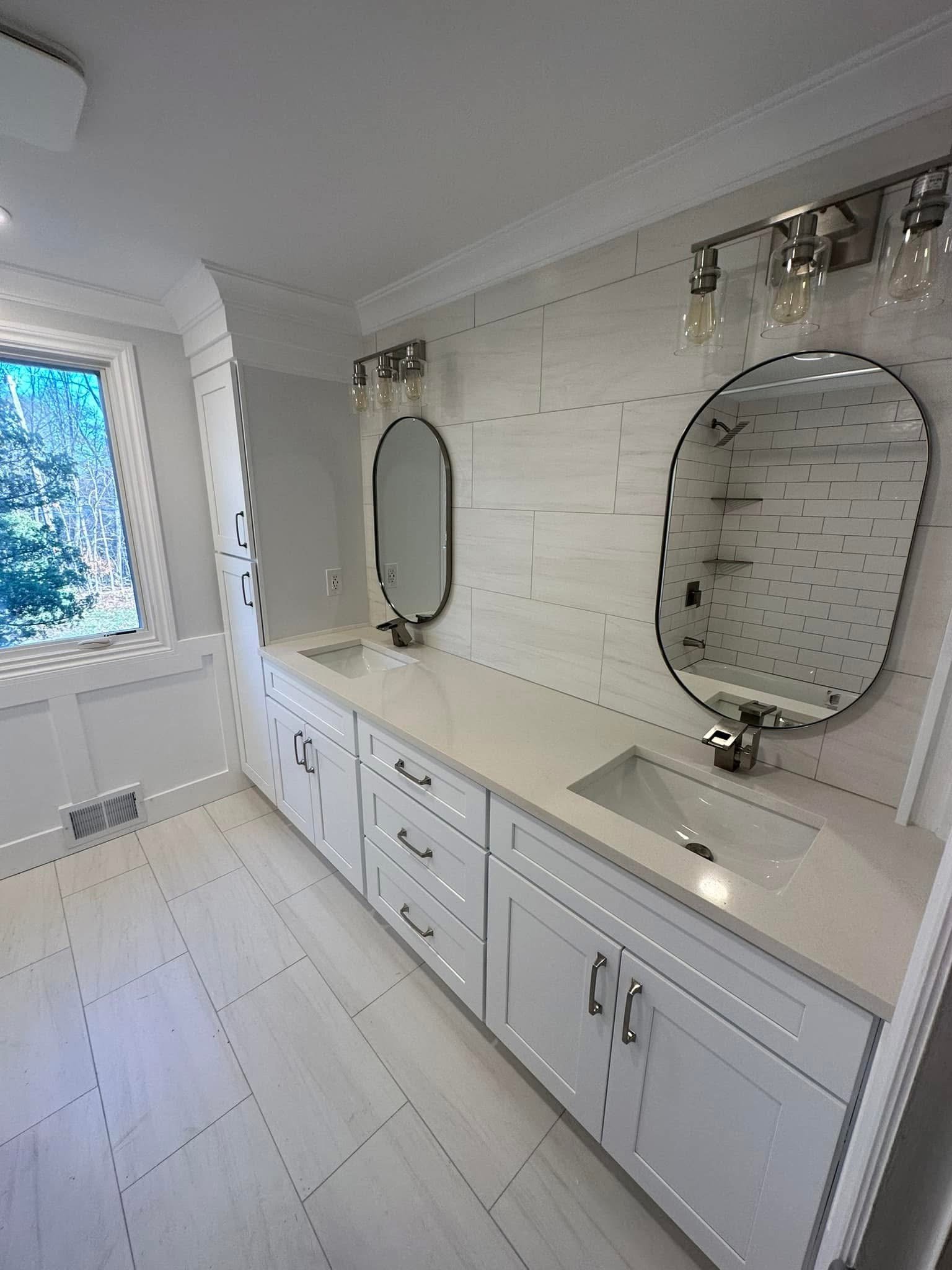 Modern bathroom with double vanity, white cabinets, two wall-mounted oval mirrors, and a white tile wall behind. A window shows a view of trees outside.