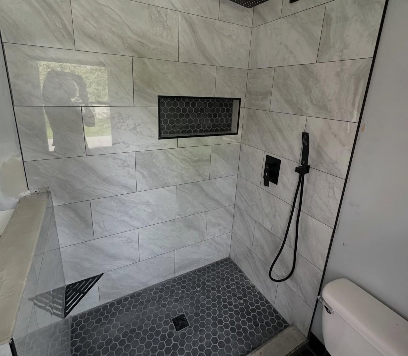 Modern walk-in shower with large rectangular marble tiles, black hexagon tiles on floor and wall niche, black handheld shower head, and white toilet in the corner.