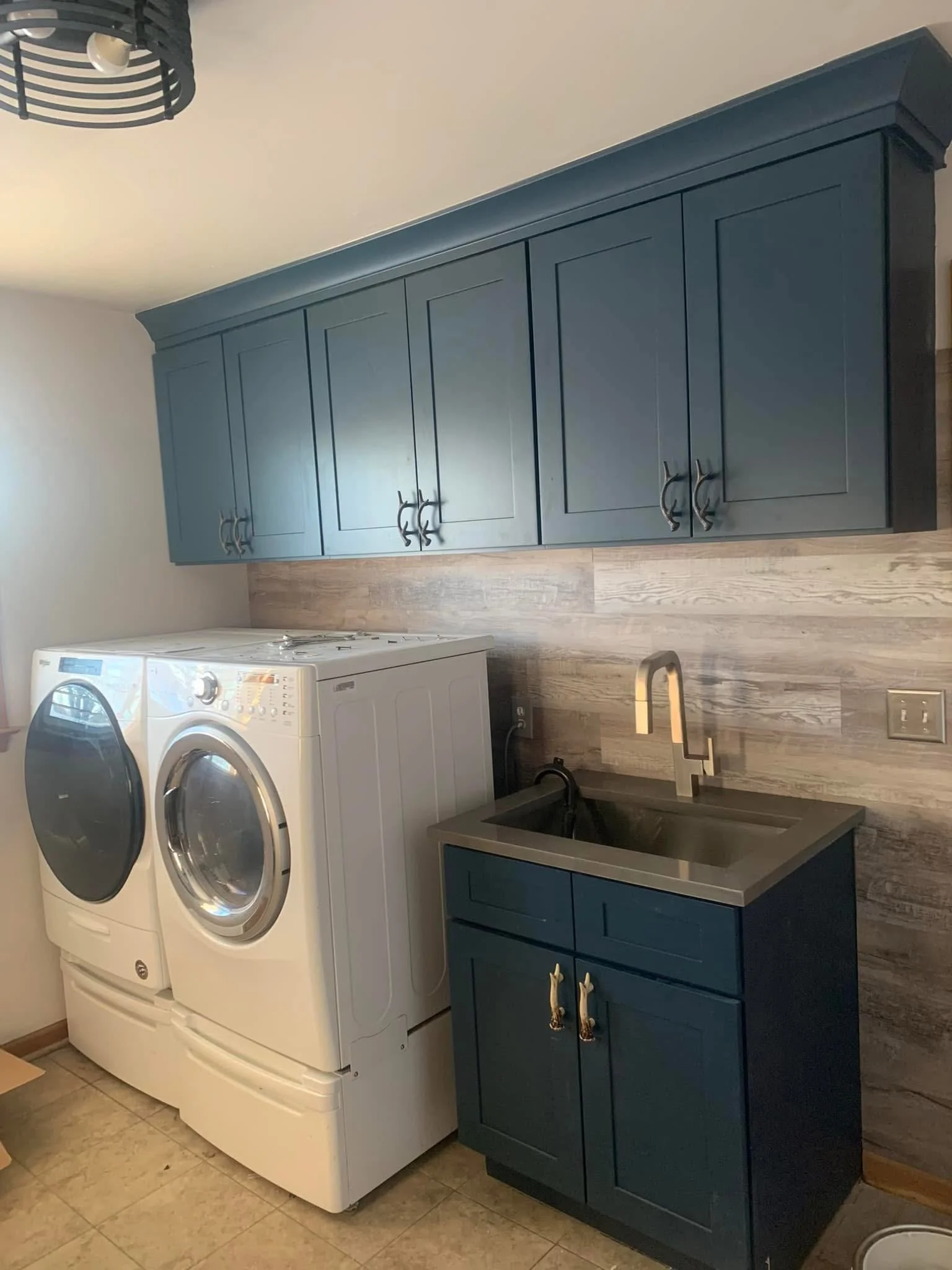 Laundry room with white washer and dryer, blue cabinets, a small sink with a silver faucet, wood-like wall paneling, and tiled flooring.