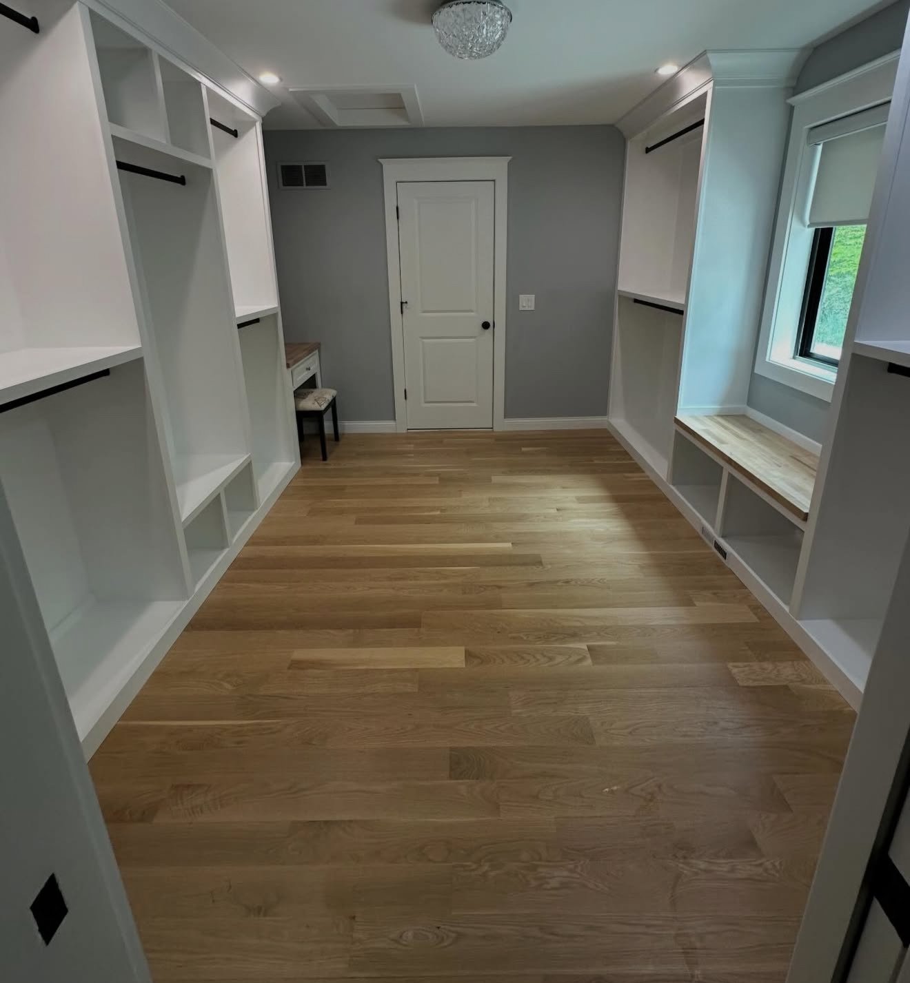 Empty walk-in closet with built-in white shelving, black clothing rods, a small desk and stool, hardwood floors, a window, and a ceiling light fixture.