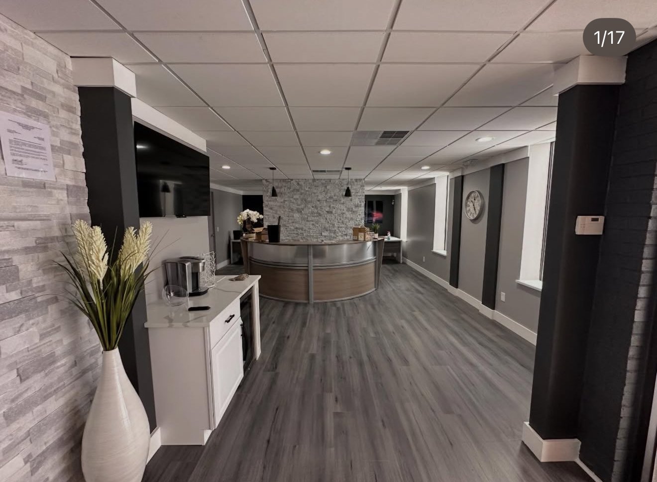 Modern reception area with a curved wooden desk, stone accent wall, gray walls with black vertical panels, large window, clock on the wall, and decorative vase with plants in the foreground.