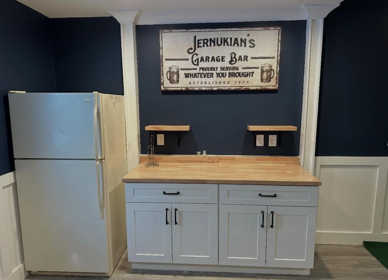 A small craft bar area with a white refrigerator on the left, a wooden countertop with a sink in the center, and a dark blue wall with a framed sign reading 'Jernukián's Garage Bar' above. There are two small floating shelves on the blue wall and multiple electrical outlets.