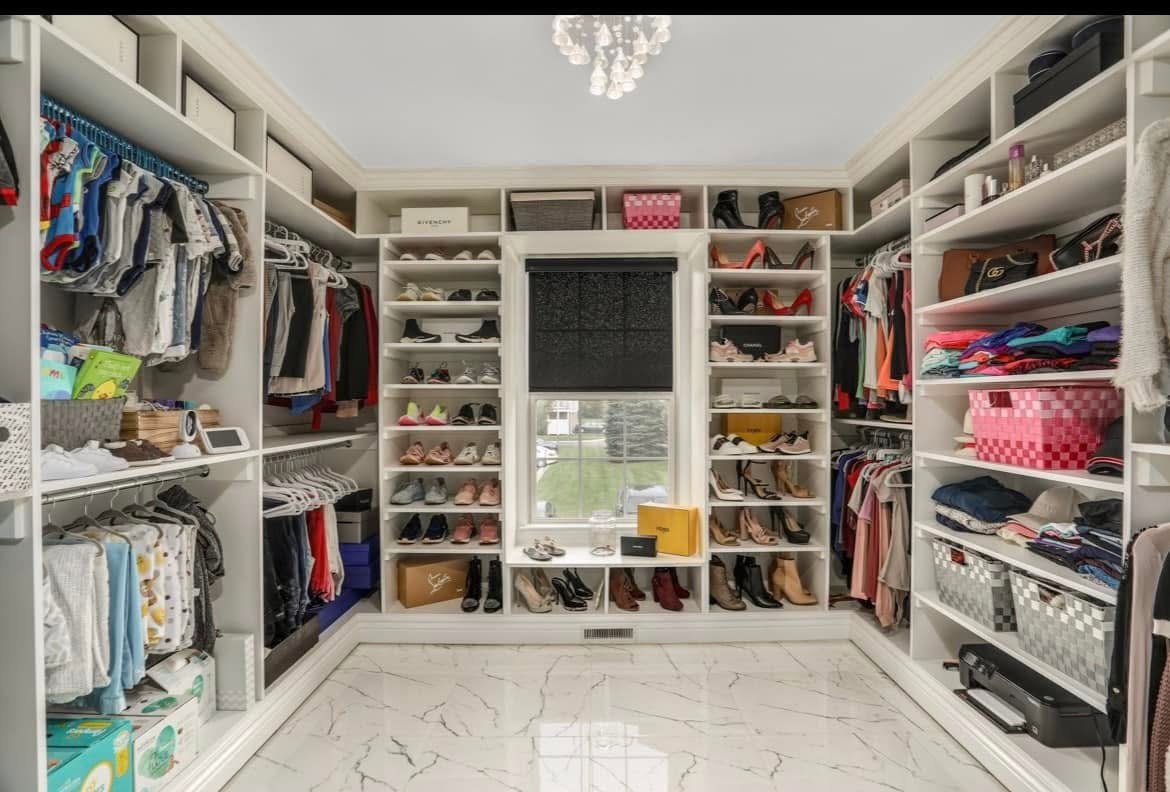 A walk-in closet with white shelves filled with clothes, shoes, and accessories. There is a window in the center with a black shade, a chandelier overhead, and a marble floor.