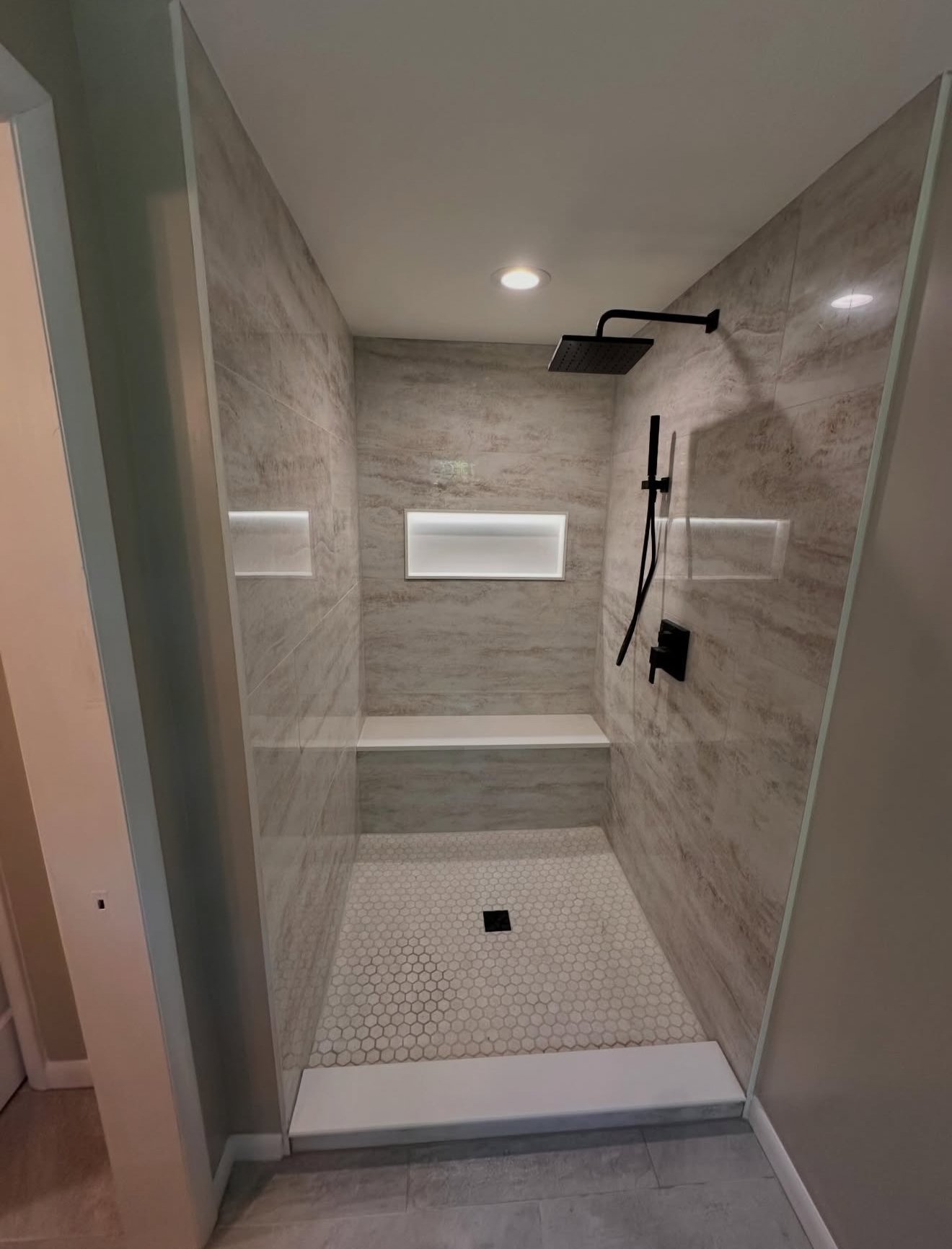 Modern walk-in shower with gray marbled tiles, black fixtures, a built-in bench, niche shelves with LED lighting, hexagonal floor tiles, and a ceiling drain.