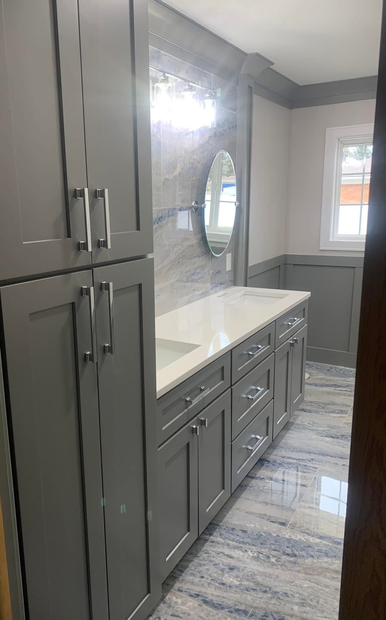 A modern bathroom with gray cabinets, a white countertop with twin sinks, a round mirror, a marble backsplash, and a window allowing natural light.
