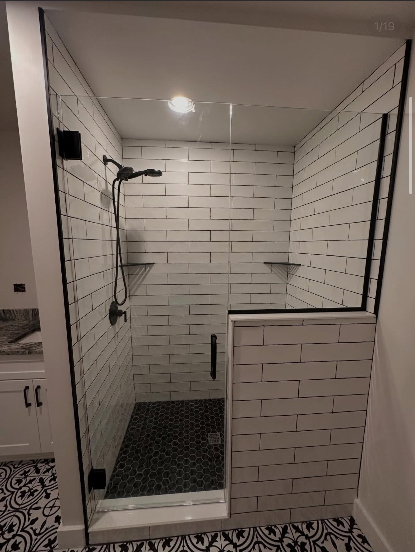 A modern walk-in shower with white subway tile walls, black hexagon floor tiles, a black shower head, and a partial glass enclosure with a black handle, inside a bathroom.