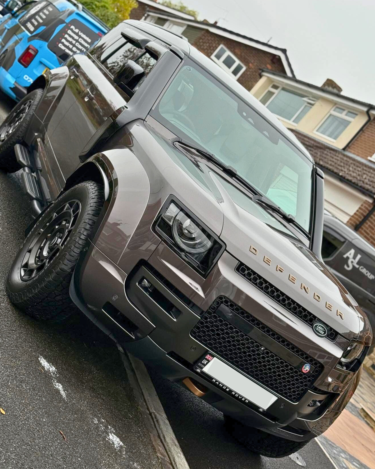 A freshly valeted black Range Rover Defender parked on a residential street with other cars and houses in the background.