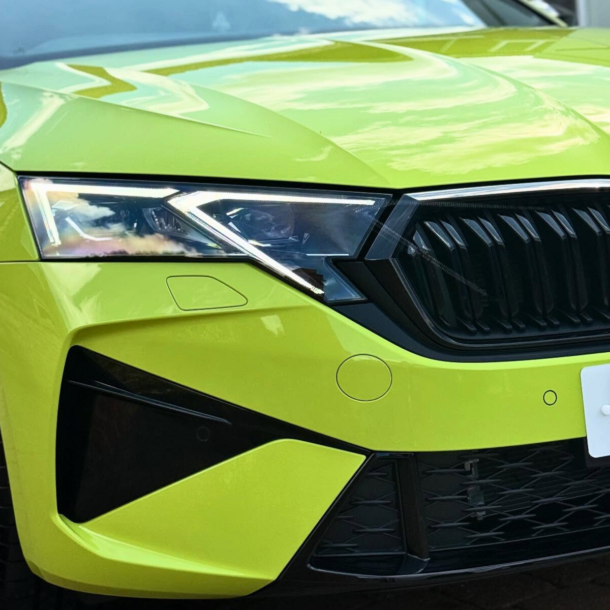 Close-up of a bright yellow car's front end, showing the gleaming paint, headlight, grille, and part of the hood and bumper.