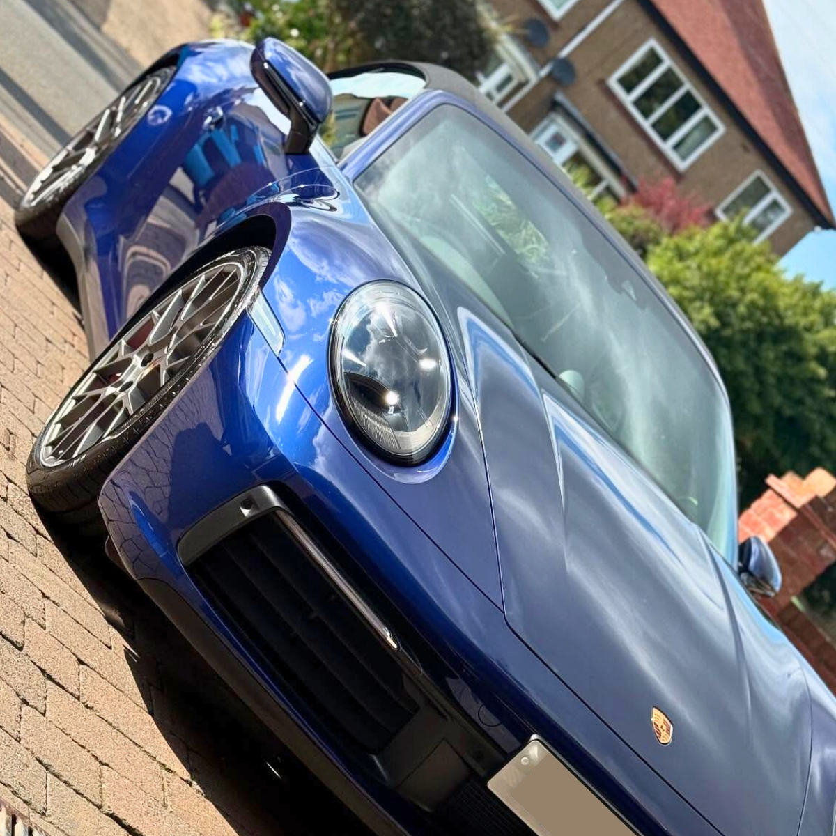 A shiny blue Porsche sports car parked on a brick driveway in front of residential houses with trees and a blue sky reflecting on the vehicle's surface.