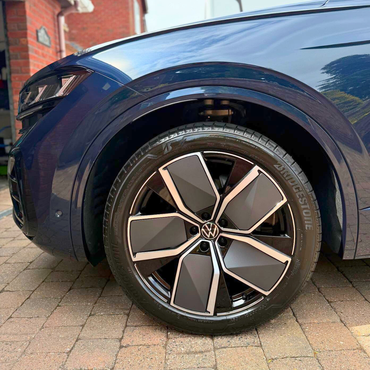 Close-up of the wheel of a shiny Volkswagen with a Bridgestone tire, parked on brick pavement outside a building.