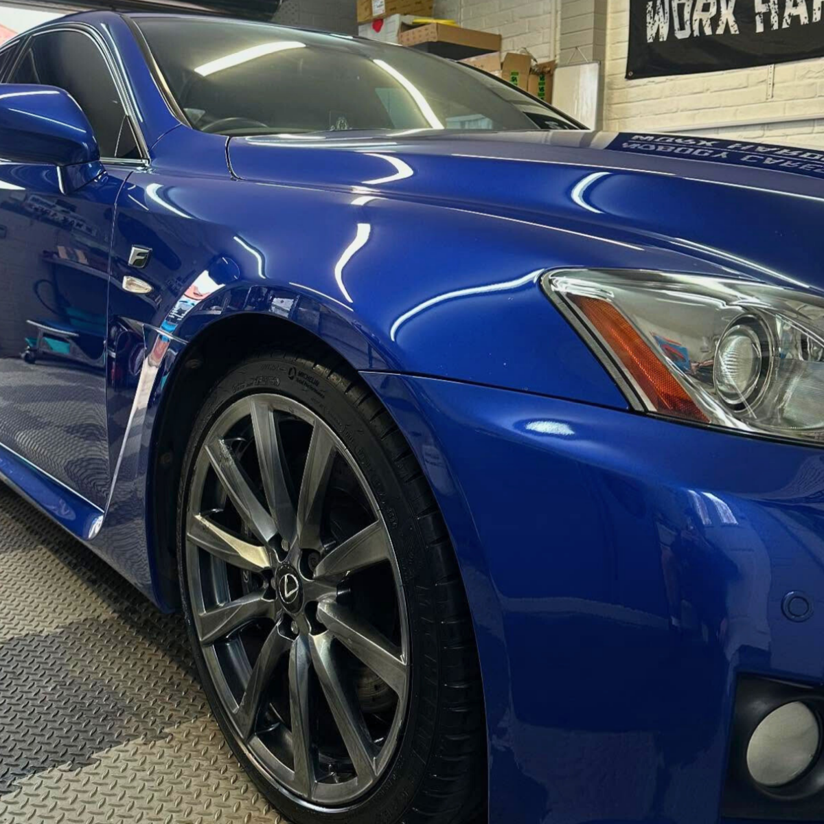 Close-up of a glossy blue sports car showing the front left side, including the tire, wheel, headlight, and part of the hood in a garage setting.