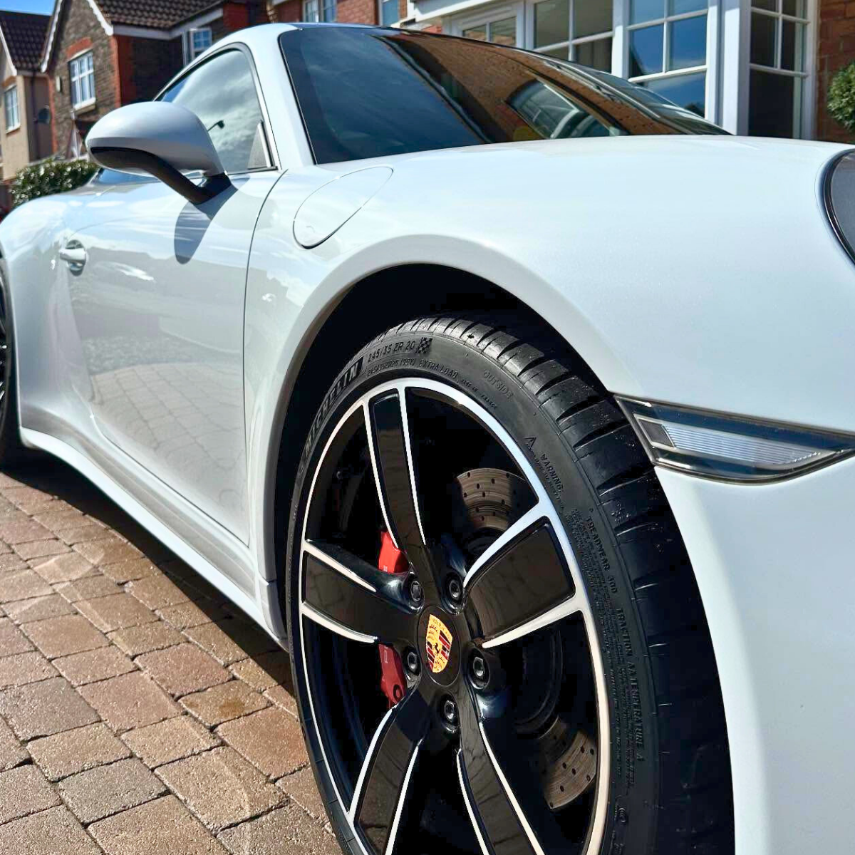 Close-up of a freshly-valeted white Porsche sports car parked on a brick driveway, showing the front wheel, tire, brake caliper, side mirror, and part of the car's body, with residential houses in the background.