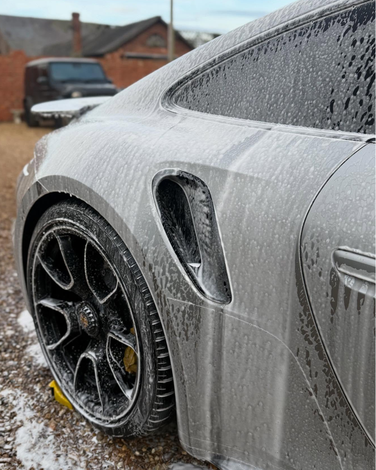 A silver car during it's detail cleaning, with soap, foam, and water, with an open wheel and a partially visible background of other vehicles and buildings.