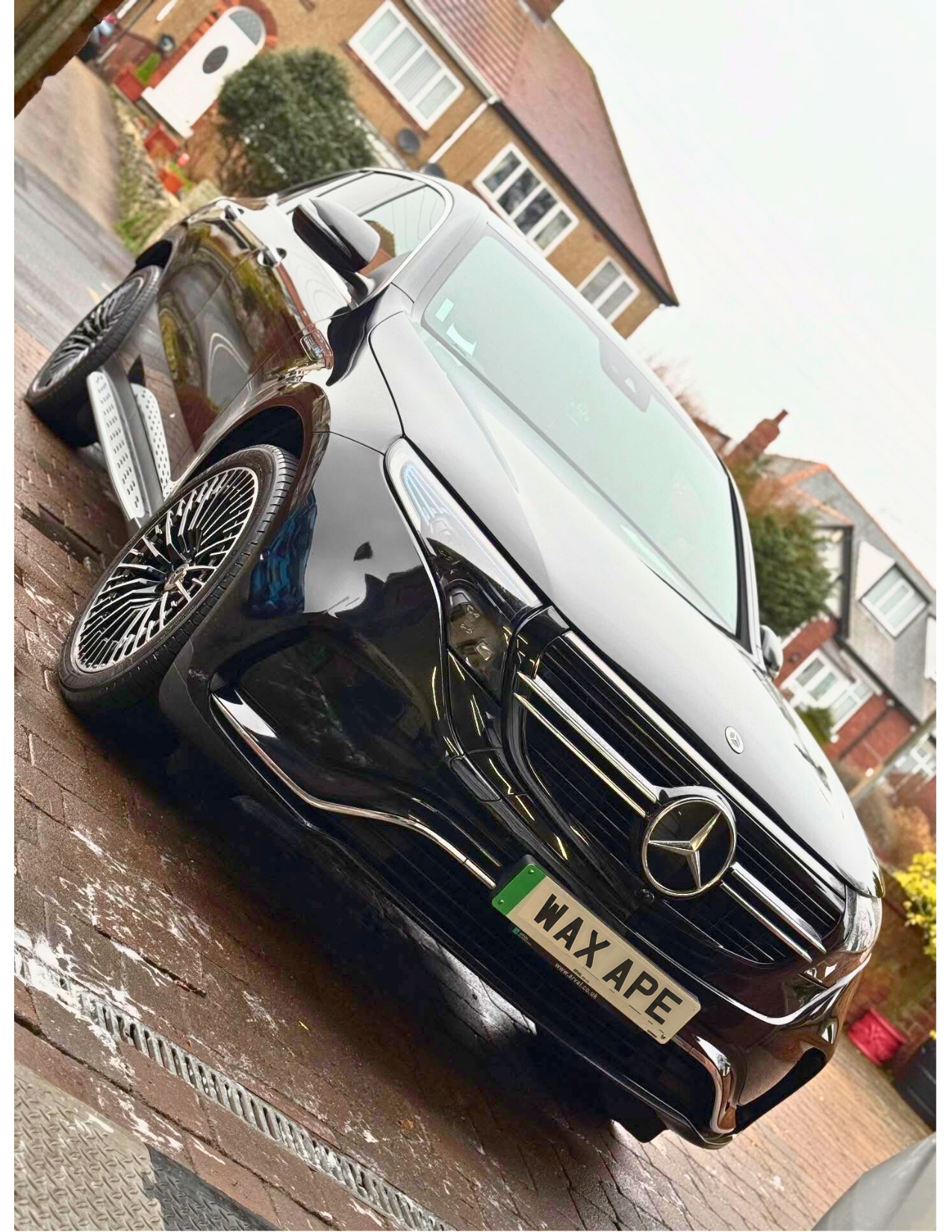 A black Mercedes-Benz parked on a brick driveway in front of a house with white-framed windows and a pointed roof. The car has an unusual license plate that reads 'WAX APE'.