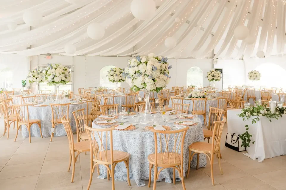 Elegant banquet hall decorated with white flowers, draped fabric ceiling, and set tables with floral centerpieces for a wedding or special event.