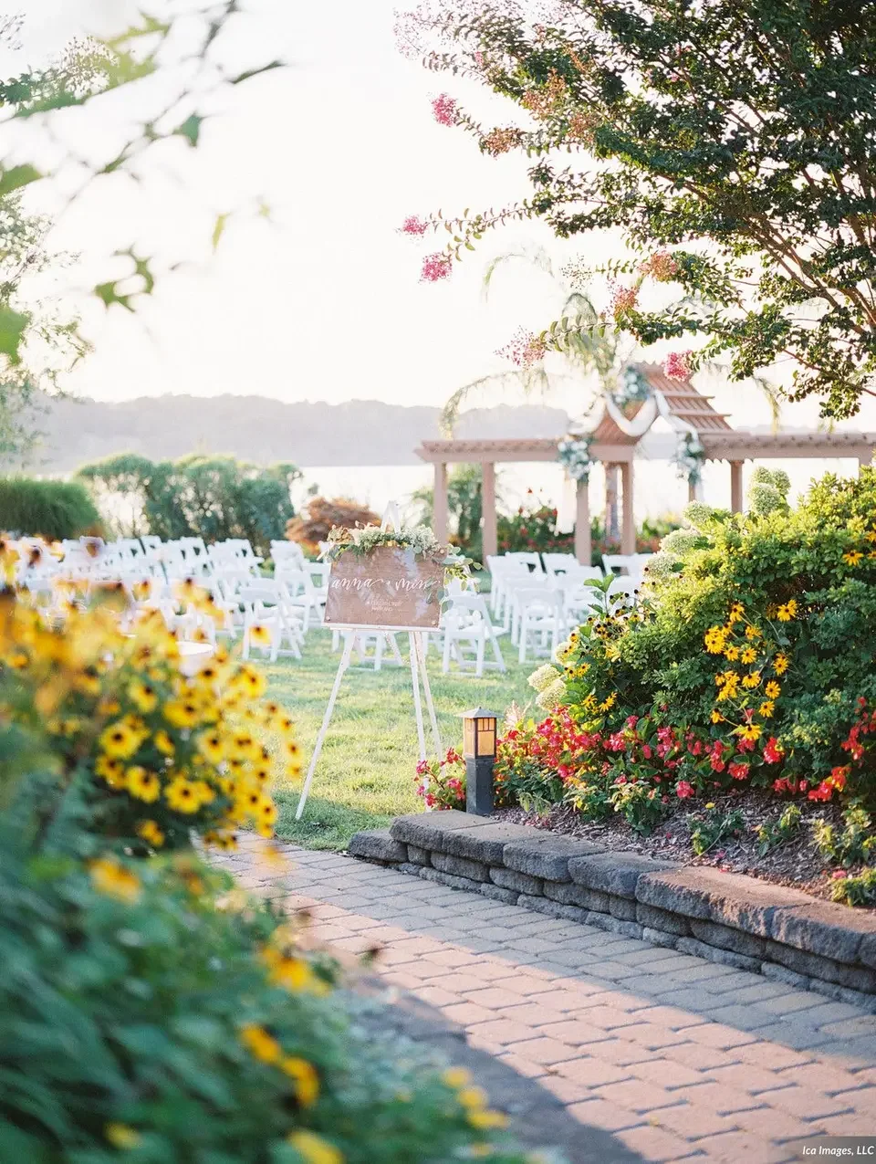 An outdoor wedding setup at sunset with white chairs arranged on a grassy area, decorated with flowers and a small pavilion in the background, surrounded by vibrant yellow and pink flowers.