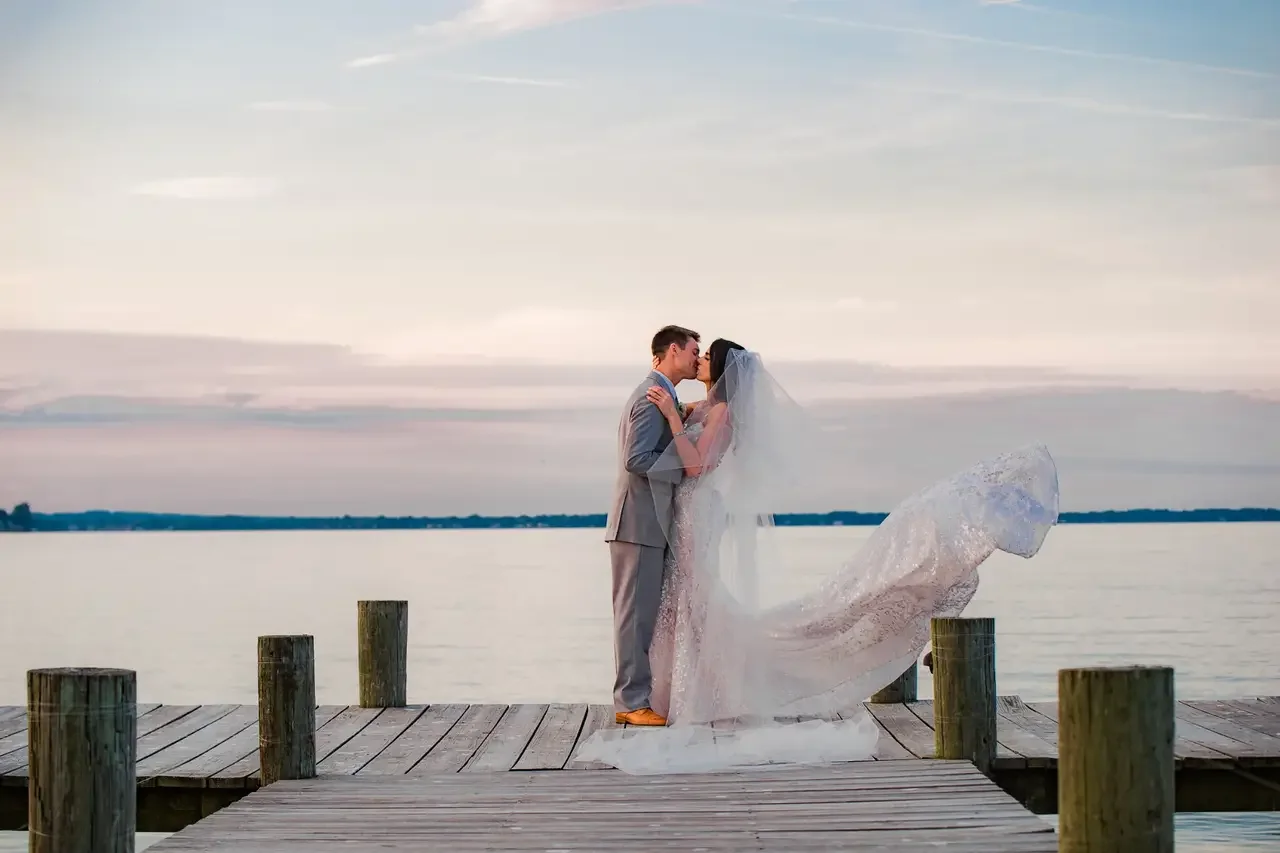 A bride and groom kissing on a wooden dock by the water during sunset, with an ice sculpture at their feet.