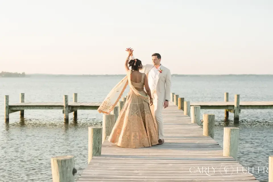 Couple dressed in formal attire standing on a wooden dock by the water during sunset, holding hands and looking at each other.