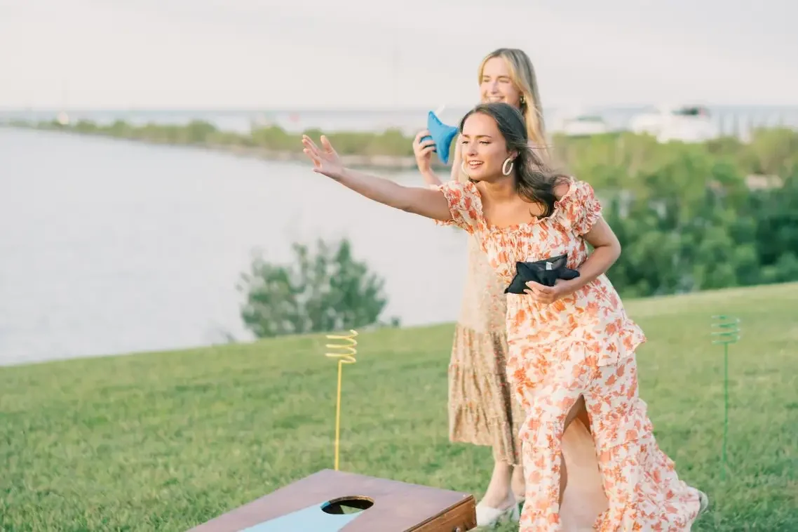 Two women outdoors near a body of water, one woman throwing a bean bag in a game, the other holding a bean bag, with a lawn and trees in the background.