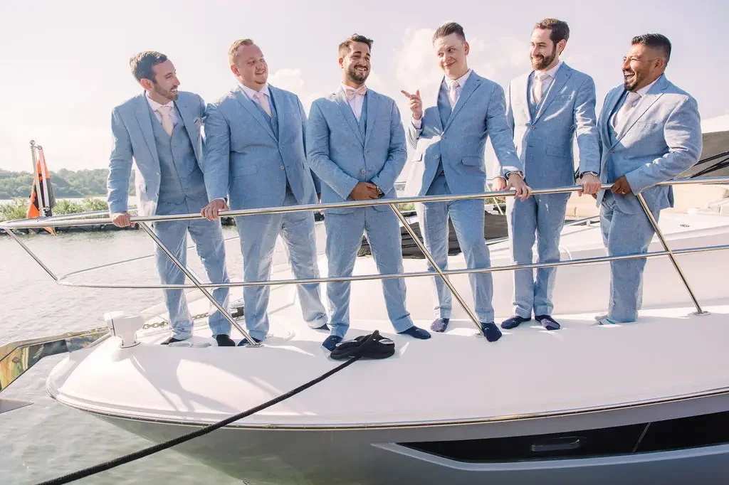 Group of seven men dressed in light blue suits standing on a boat, engaging in conversation, near a water body with trees and a partly cloudy sky in the background.