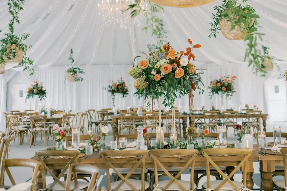 Elegant wedding reception setup with long wooden tables, floral centerpieces, and hanging greenery in a white tent.