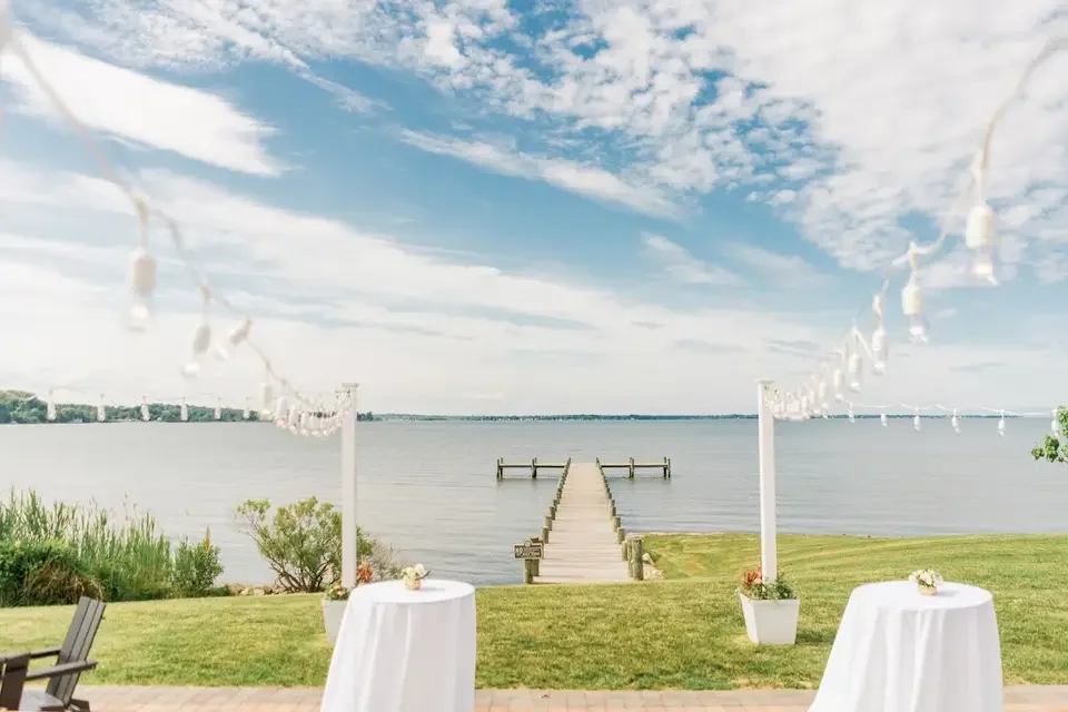 A lakeside outdoor setting with two round tables covered in white cloths, small flower arrangements, and potted plants, under string lights. A wooden dock extends into a calm lake under a partly cloudy sky.