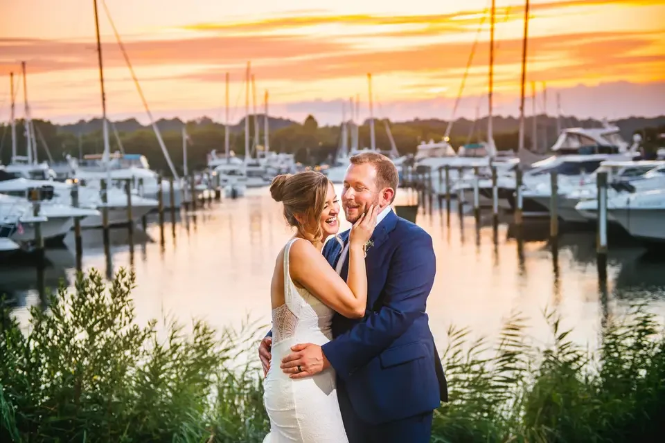 A newlywed couple is embracing and smiling by a marina at sunset, with yachts docked in the background.